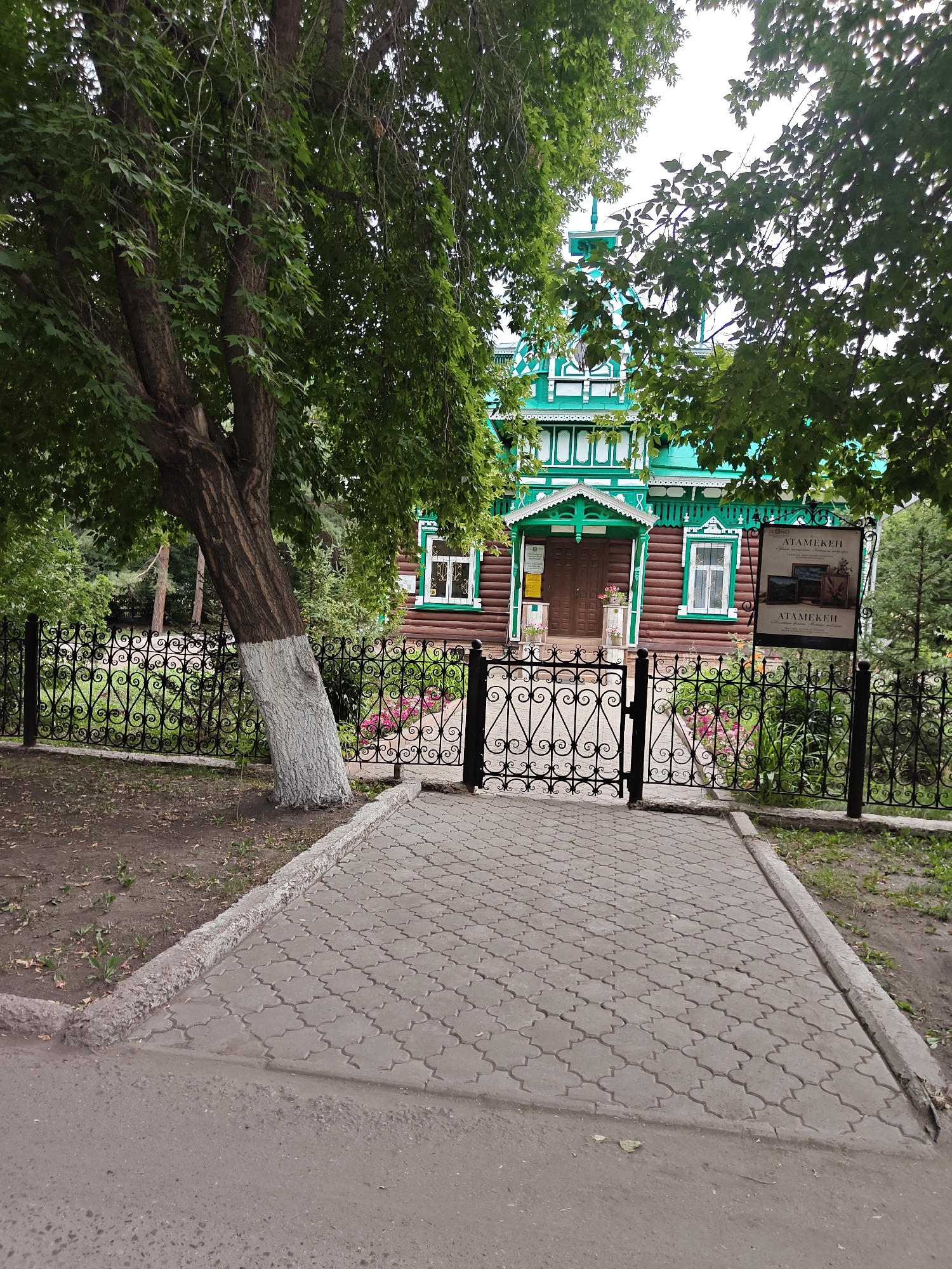 The image shows the exterior of the Abai House Museum in Petropavl, Kazakhstan. The museum is a traditional wooden house painted red and green, with intricate white trim. It is surrounded by a black wrought-iron fence and a paved walkway. A large tree with white-painted trunk stands to the left of the walkway. A sign with the text "АТАМЕКЕН" (Atameken) is visible to the right of the entrance. The weather appears overcast.