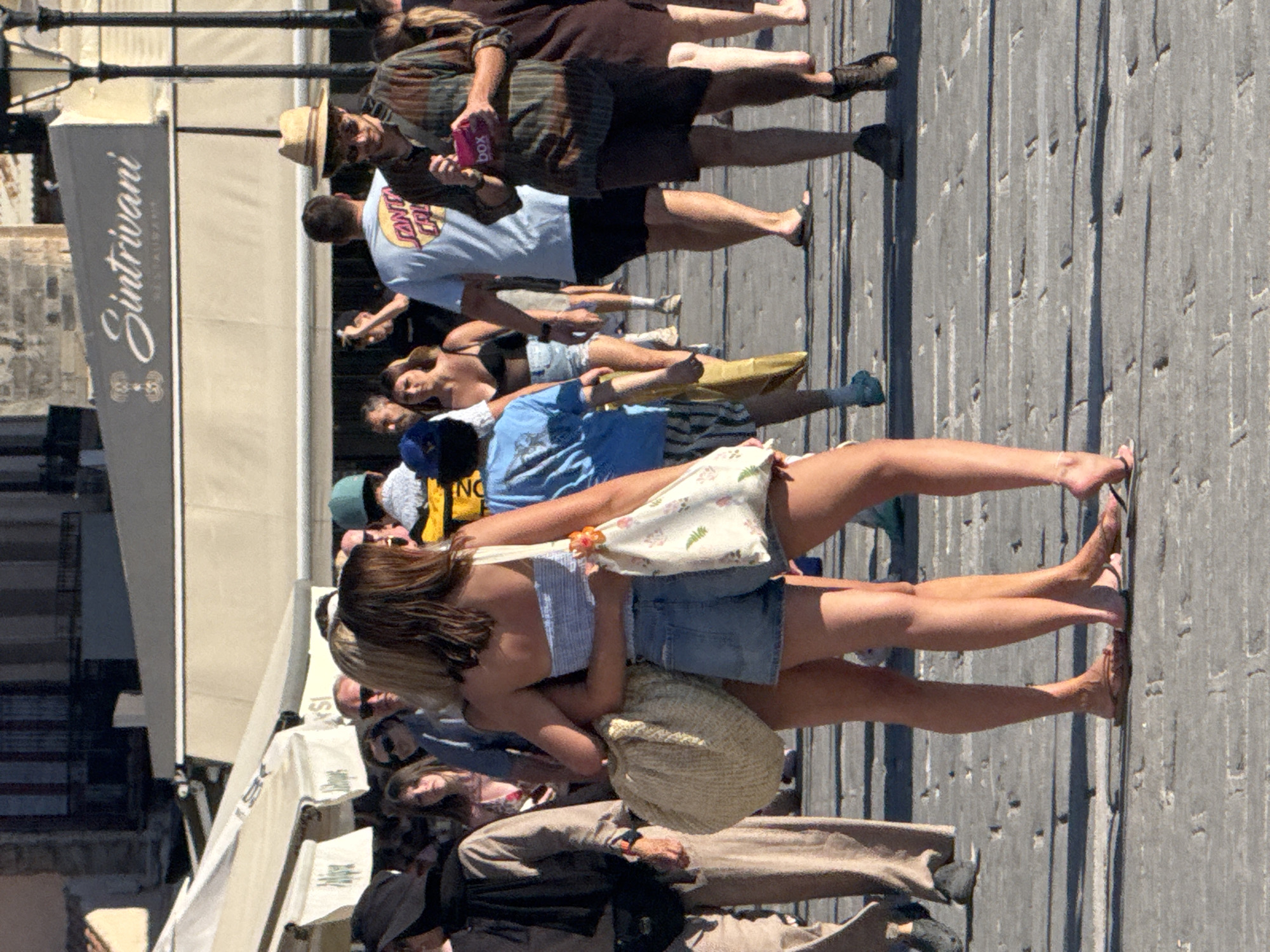 In Chania, Greece, on a paved promenade, two young women are embracing. The woman on the left has blonde hair and is wearing a light blue strapless top and denim shorts. The woman on the right has brown hair and is wearing a white and floral print top. She is also wearing denim shorts. Both women are carrying large tote bags. They are surrounded by a crowd of people walking in both directions. The background shows buildings with awnings and shops. In the distance, a sign reads "Sintrivani Restaurant". The scene is brightly lit, suggesting it is daytime.