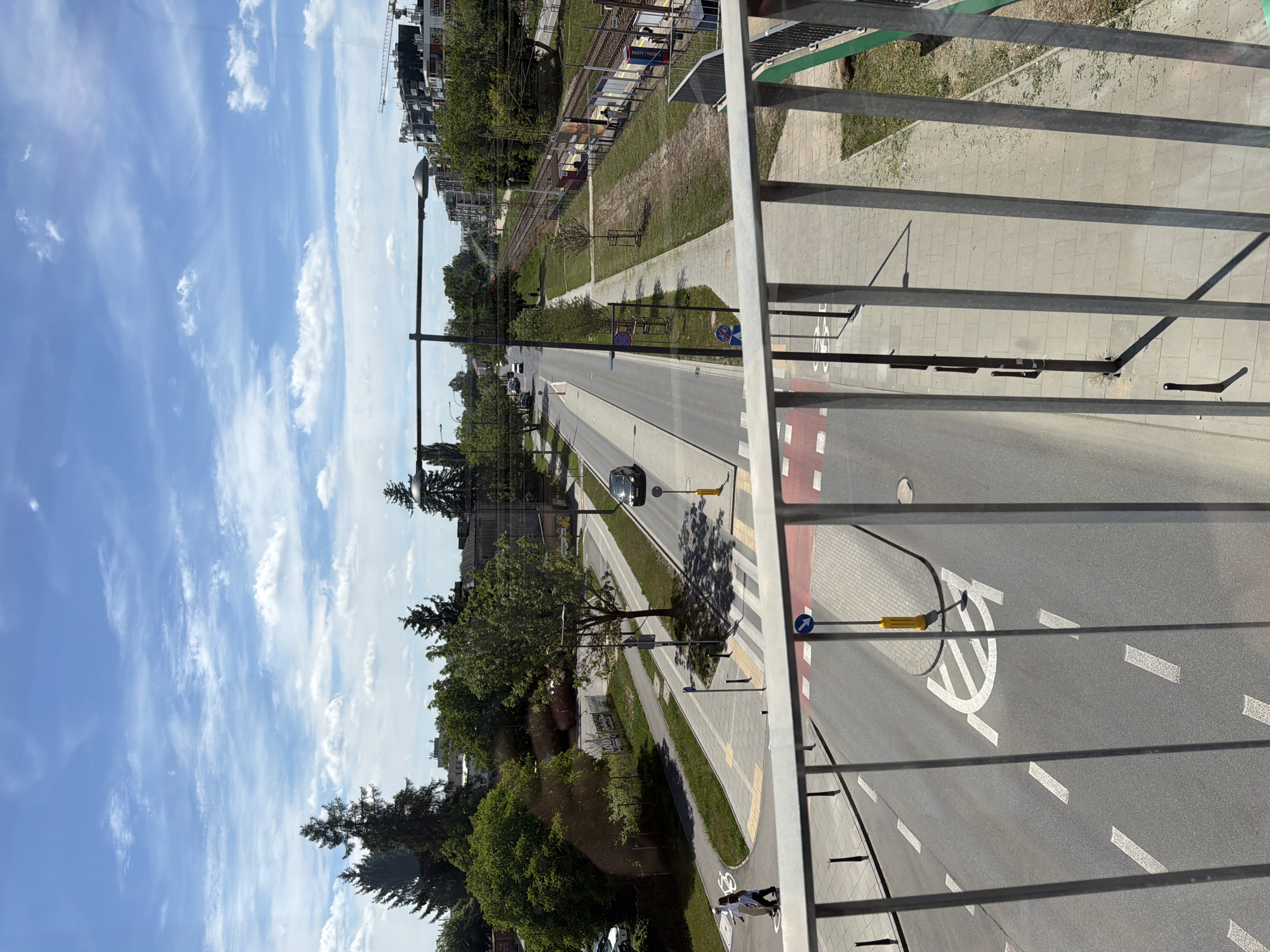 A high-angle, full shot captures a street scene with a pedestrian bridge in the foreground. The bridge's metal railing runs horizontally across the bottom of the frame. Below, a two-lane road with white lane markings and a pedestrian crossing is visible. A pedestrian crossing light pole stands at the crossing. To the right of the road, a pedestrian walkway is bordered by a grassy area and railroad tracks. On the far right, a train platform with people and a train is partially visible. Beyond the tracks, modern buildings and construction cranes indicate an urban environment. The sky is blue with scattered white clouds. Trees and other greenery are interspersed throughout the scene.