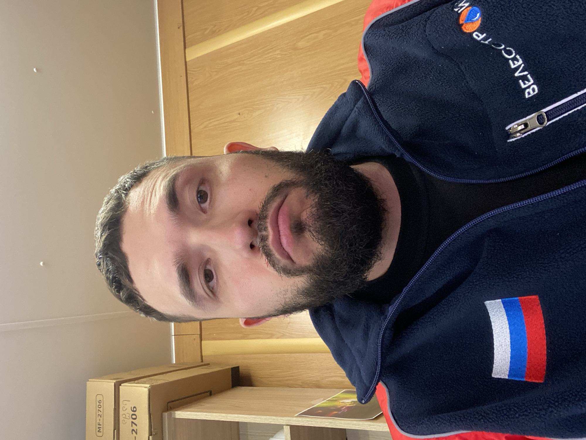 A man with a beard is looking at the camera. He is wearing a navy blue jacket with a white, blue, and red Russian flag patch on the lower left side. The jacket also has the word "ВЯЗАССЕТРОЙ" embroidered on the upper right side with a globe image above it. The background includes a wooden wall and shelving unit. There are cardboard boxes on one of the shelves. This image does not provide enough context to confirm a location in Luzhitsy, Russia.
