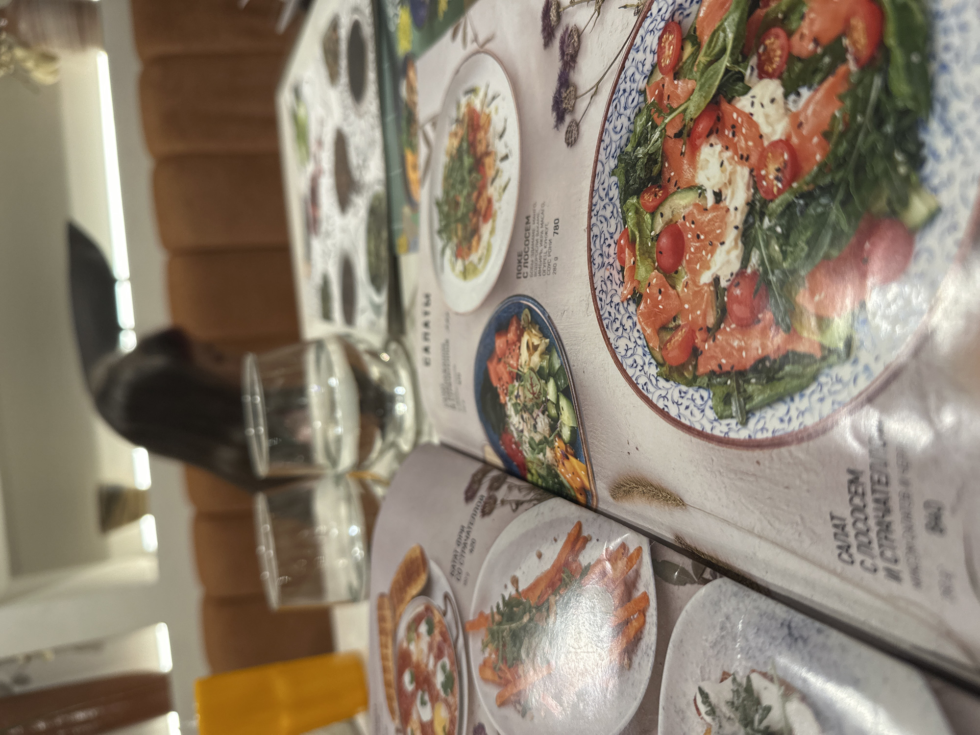The image shows a close-up view of a menu at a restaurant, with two glasses of water and an orange bottle visible in the background. The menu features pictures and descriptions of various dishes, including salads and what appears to be a breakfast item. One of the salads prominently displayed is a salmon salad with tomatoes, avocado, and greens. The menu is written in Russian. The restaurant appears to be moderately lit, with orange upholstered seating in the background. There is no explicit indication of the city or country.