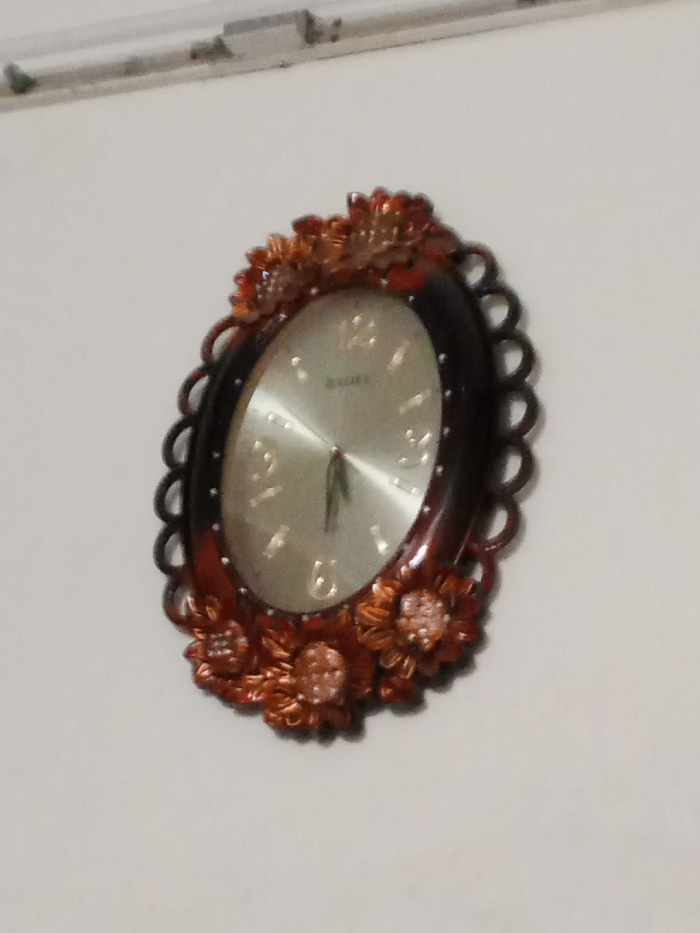 The image shows an oval, wall-mounted clock with a brown wooden frame. The frame has a decorative floral design around the edge. The clock's face is silver with Roman numerals, and the hands are black. The clock is mounted on a white wall. It appears to be hanging in a residential location in Sialkot, Pakistan.