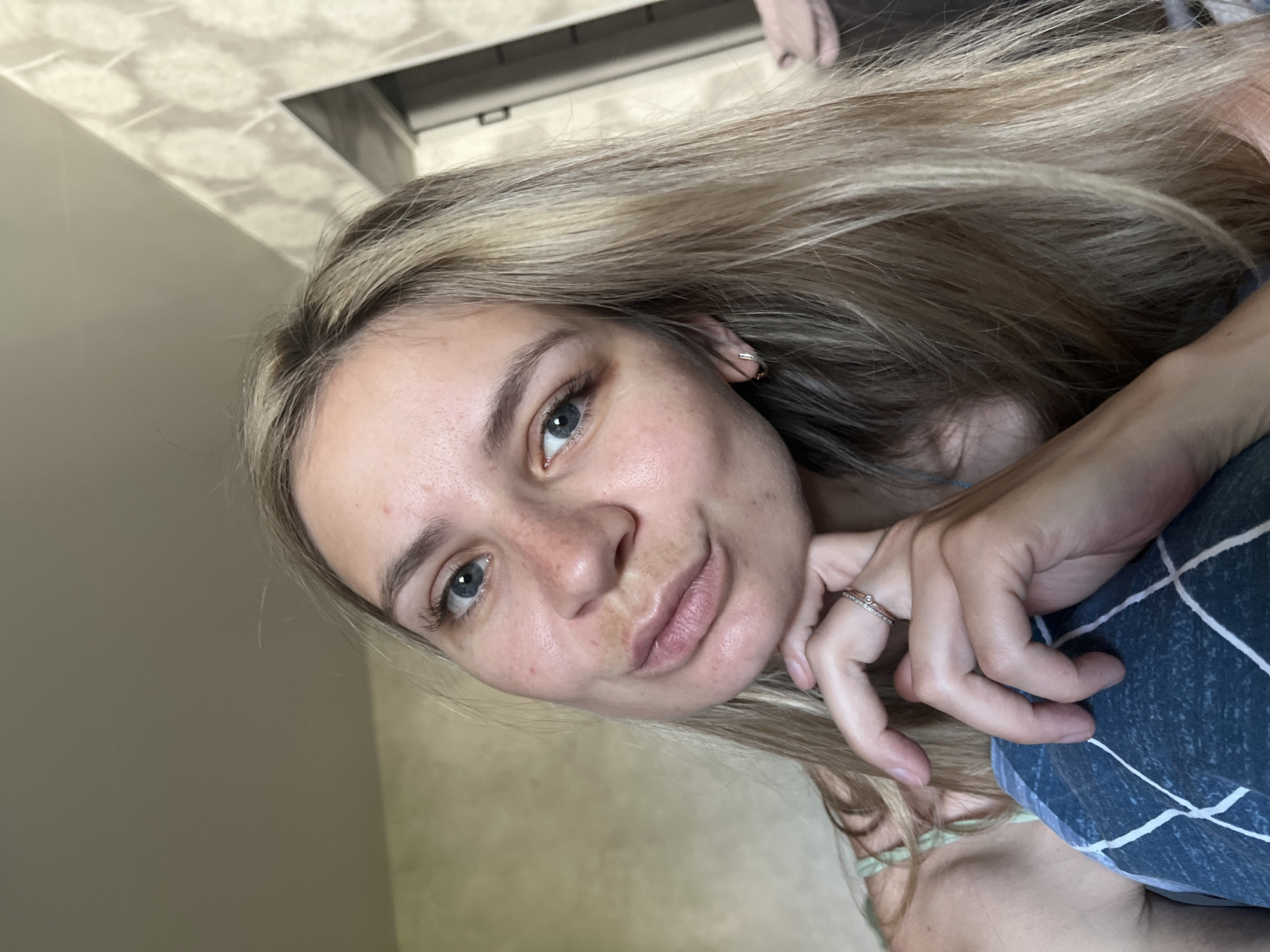 A close-up selfie features a woman with blonde hair and blue eyes, posing indoors. She is wearing a ring on her finger and is resting her face on her hand. The background is slightly blurred, showing a wall and a glimpse of a ceiling fixture. The photo appears to be taken at an upward angle. No information on the city of Chaikovsky, Russia, can be extracted from the image.