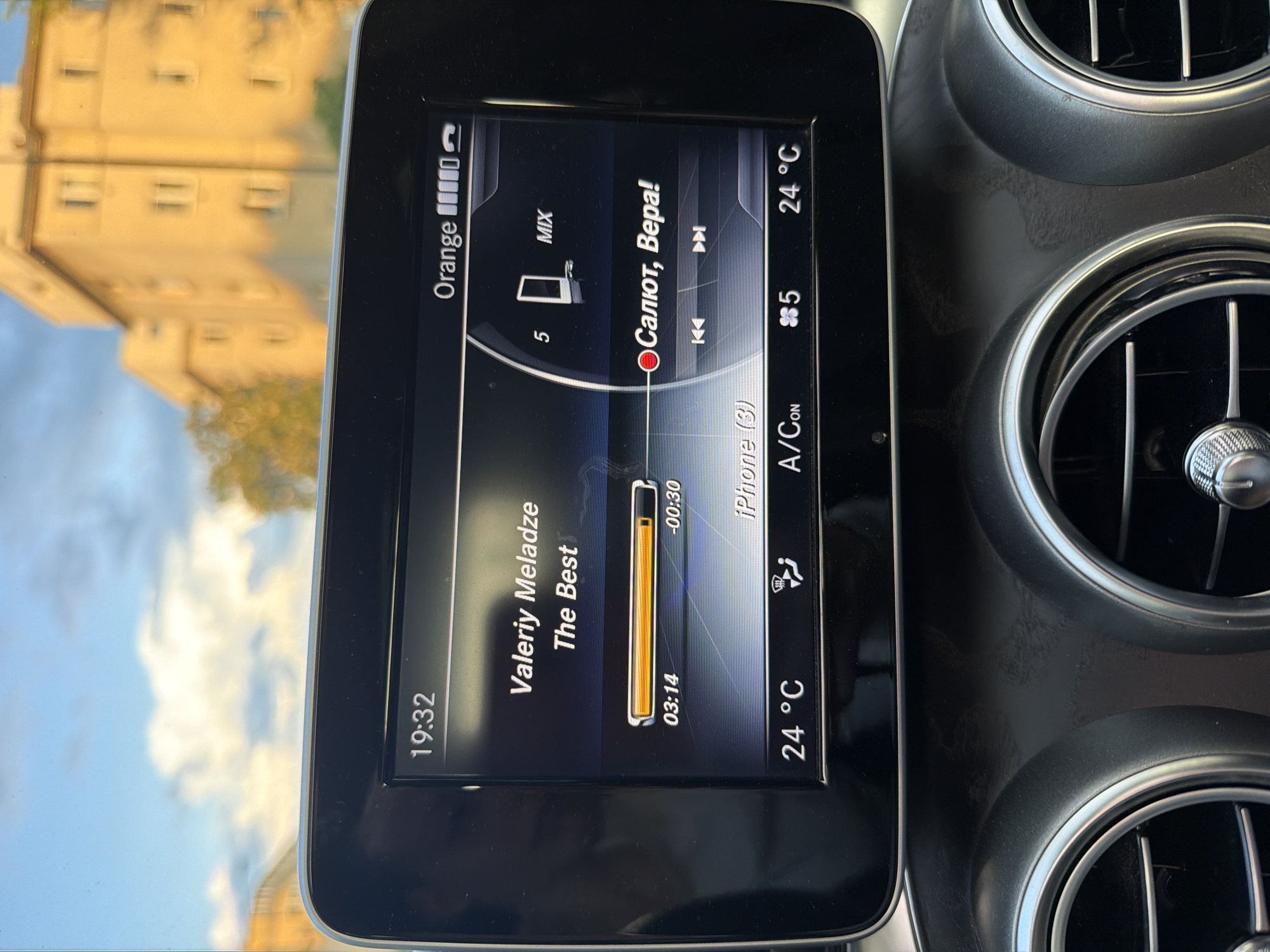 The image shows the dashboard of a car. The infotainment system is displaying music information: the artist is Valeriy Meladze, the song is "The Best", playing from an iPhone (3). The next song is "Salut, Vera". The time is 19:32, the temperature is 24 degrees Celsius, and the air conditioning is on. The network is "Orange" and the signal strength is full. Part of a building and sky are visible reflected in the screen. The city and location cannot be determined from the image.