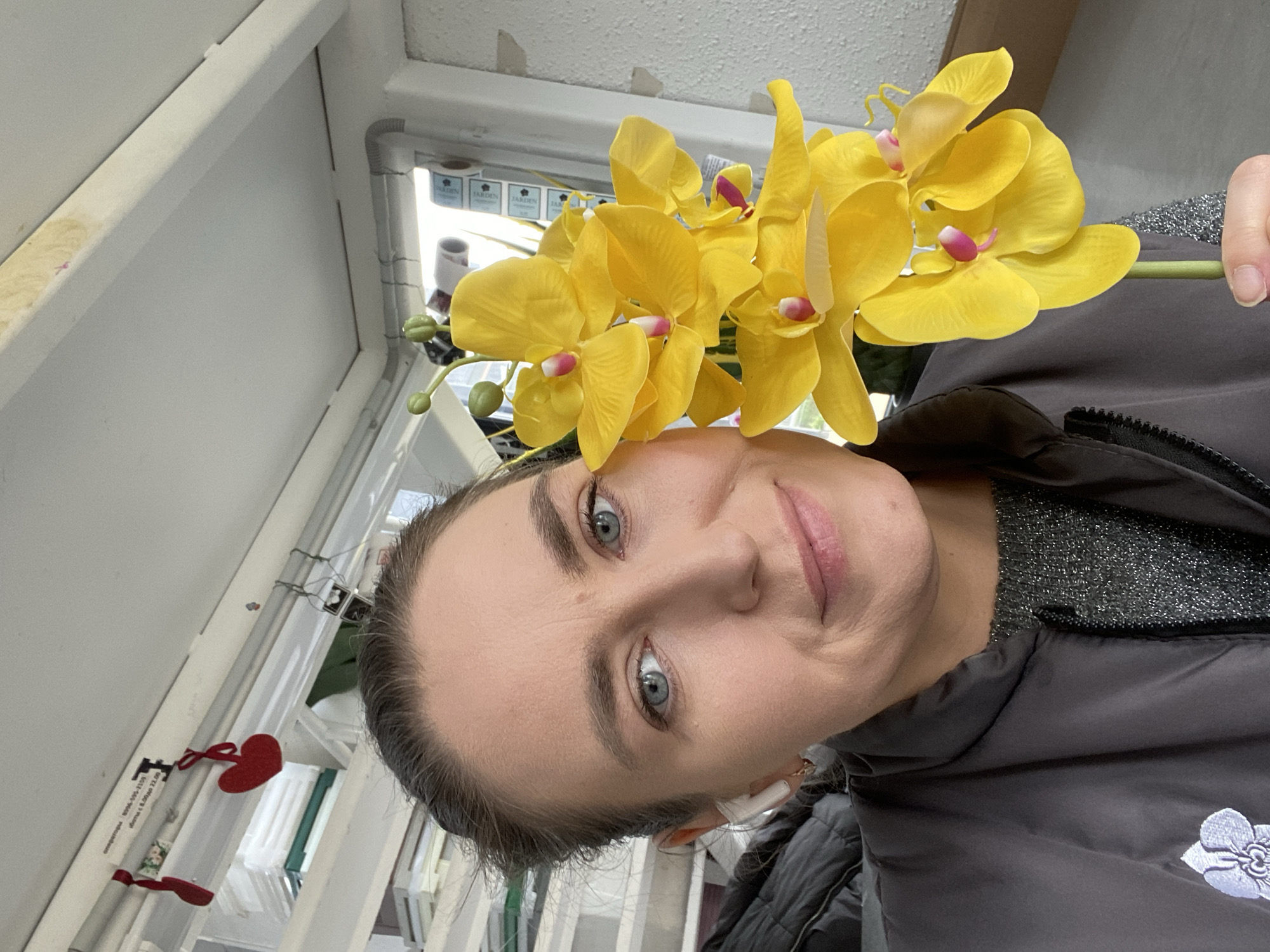 A woman with gray eyes and fair skin is holding a bunch of yellow orchids up to her head. She is wearing a dark gray jacket over a sparkly gray top. She has headphones on. In the background, there are windows, a white wall, and a door with a sign. Red heart-shaped decorations are hanging on the window frame. It is unclear if the image was taken in Yekaterinburg, Russia.