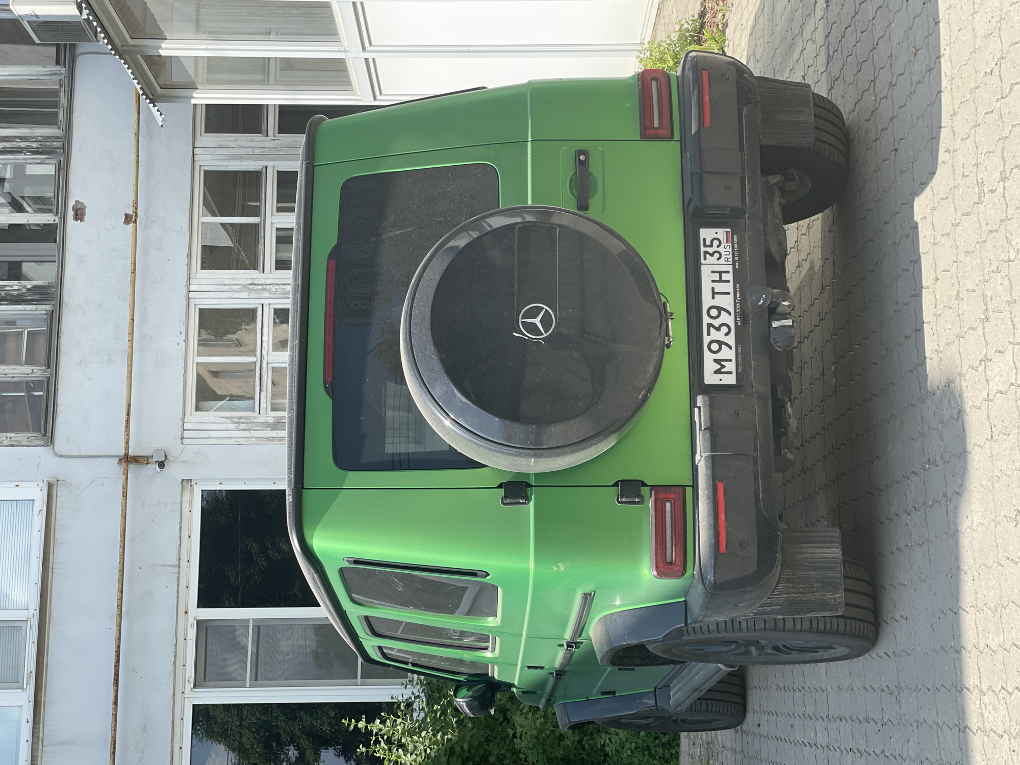 A bright green Mercedes-Benz G-Class SUV is parked on a paved surface in front of a light-colored building. The SUV has a spare tire mounted on its rear and a Russian license plate reading "M939TH 35 RUS". The building appears to be an industrial or commercial structure with multiple windows and some signs of wear. Green bushes are visible to the left of the vehicle. The overall scene depicts a parked vehicle in an urban or semi-urban environment.