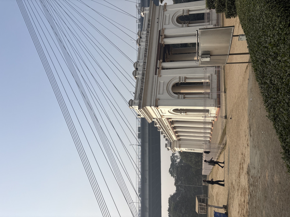 A white, ornate building with arched windows and columns stands adjacent to a suspension bridge with numerous support cables. The structure is bordered by a green hedge and a sandy path where several people are walking. Tall trees are visible in the background under a clear, pale sky. A blue trash receptacle sits at the edge of the pathway. The scene captures an architectural landscape under a bright, overcast sky.