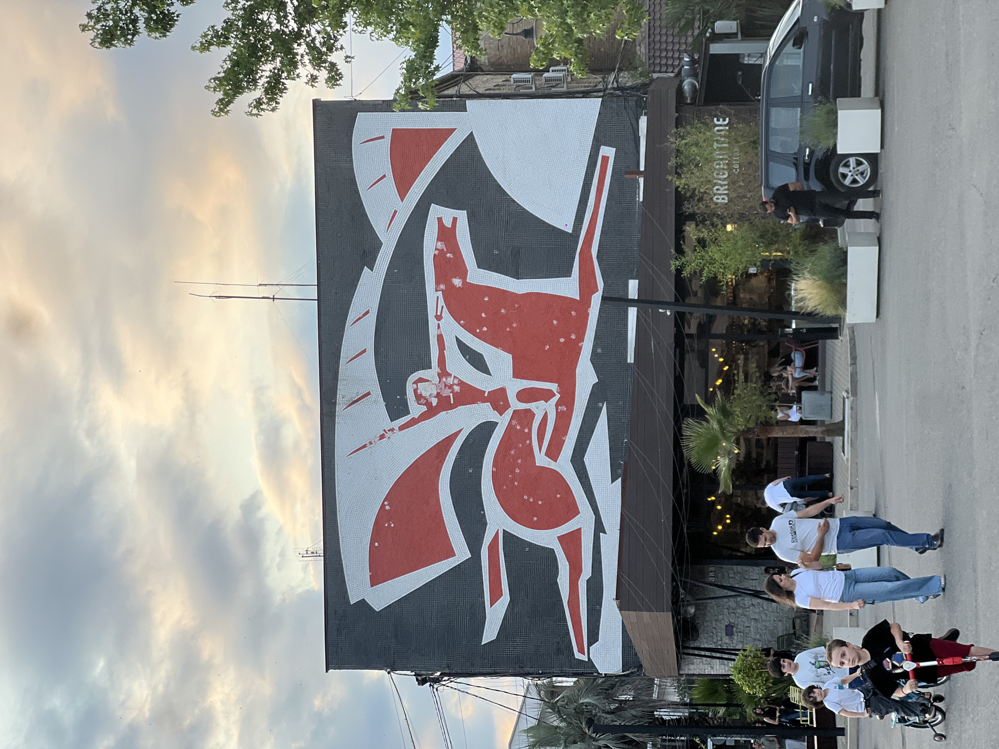 The image shows a street scene with a large mosaic mural on the side of a building. The mural is abstract, featuring bold red and white geometric shapes on a dark background, with a depiction of a stylized animal or figure. Trees with green leaves overhang the top of the image, and a cloudy sky is visible.
In front of the building, a black van is parked next to a cafe with outdoor seating. The cafe has the text "BRYGHTIDE COFFEE" visible on its awning. Several people are walking on the sidewalk and street. A group of people in casual attire, including white t-shirts and jeans, are standing and walking. In the foreground, a child is sitting on a tricycle.
The scene appears to be taking place in the late afternoon or early evening, indicated by the soft lighting and the warm tones in the clouds. The weather seems to be mild. The setting is likely an urban or semi-urban area with businesses and pedestrian activity. The mood is casual and relaxed.