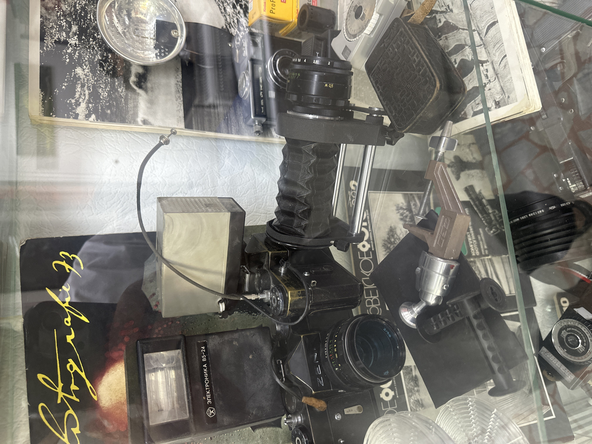 This is a photograph of vintage photography equipment displayed inside a glass case, likely in a museum or antique shop. The display includes several cameras, lenses, a flash unit, and other accessories. A book with the title "Fotografiia 73" in stylized yellow script is visible at the top of the display. In the background, a black and white photograph of a stormy sea or landscape is partially visible. The lighting appears to be artificial, casting reflections on the glass. The overall scene suggests a curated collection of historical photographic items. The location of this display is in Tobolsk, Russia, as indicated by the museum setting. There are no people visible in the photograph.