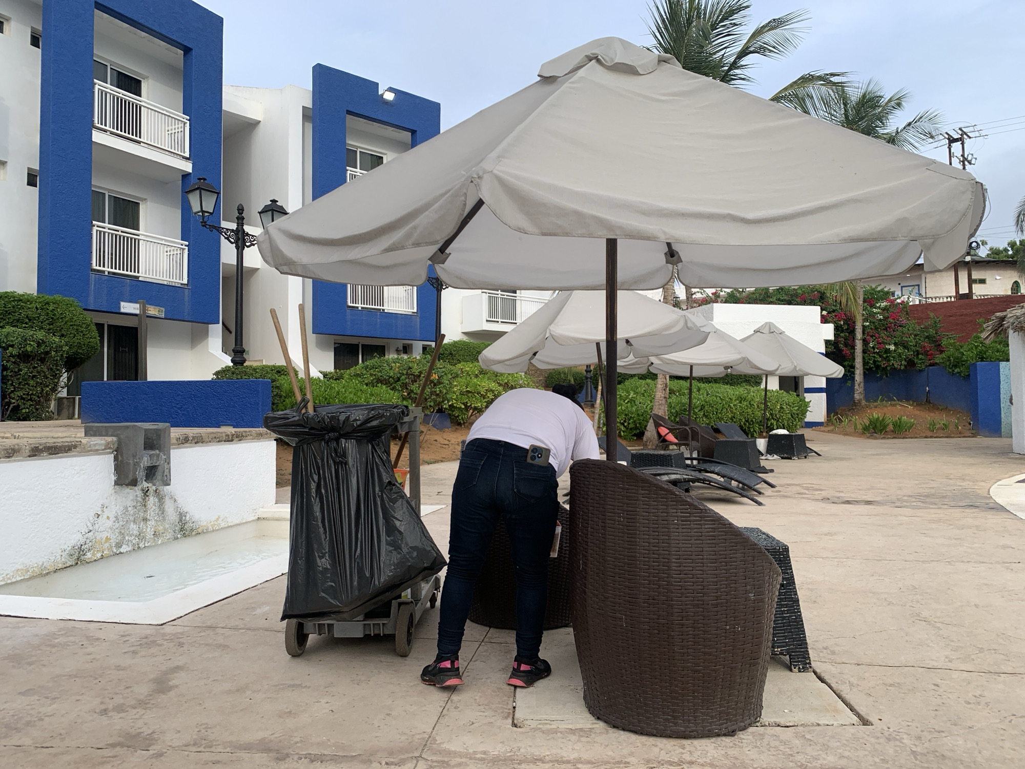 The image depicts an outdoor area, identified as a resort or hotel property in Playa El Agua, Venezuela, under an overcast sky. In the foreground, a person is observed from behind, bent over an unseen object, wearing a white short-sleeved t-shirt, dark blue jeans, and black athletic shoes with pink accents. A smartphone is tucked into the back right pocket of the jeans, and a red tag or label hangs from a belt loop. The person is positioned adjacent to a dark brown, wicker-patterned outdoor chair. To the person's immediate left stands a grey utility cart carrying a large, black trash bag. A long handle, possibly for a cleaning tool, extends upward from behind the cart.
On the left side of the foreground, a white, textured concrete wall bounds a long, shallow white basin containing water. A grey concrete spout is affixed to the wall, below which dark green and yellow staining indicates persistent water flow and algae growth.
The mid-ground is dominated by several large, off-white fabric patio umbrellas, open and providing shade. In the background, two multi-story buildings feature white exteriors with distinctive blue rectangular frames around multiple balconies equipped with white railings. A black decorative street lamp with two lanterns is positioned in front of the left building, beneath which a directional sign is visible on the building wall. Green shrubs and bushes are planted around the base of the buildings and along the edges of the paved concrete ground.
Further to the right and in the distant background, palm trees, flowering bushes with red and white blossoms, utility poles with power lines, and additional light-colored structures are discernible against the grey sky. Several dark-colored outdoor lounge chairs are scattered beyond the immediate work area.