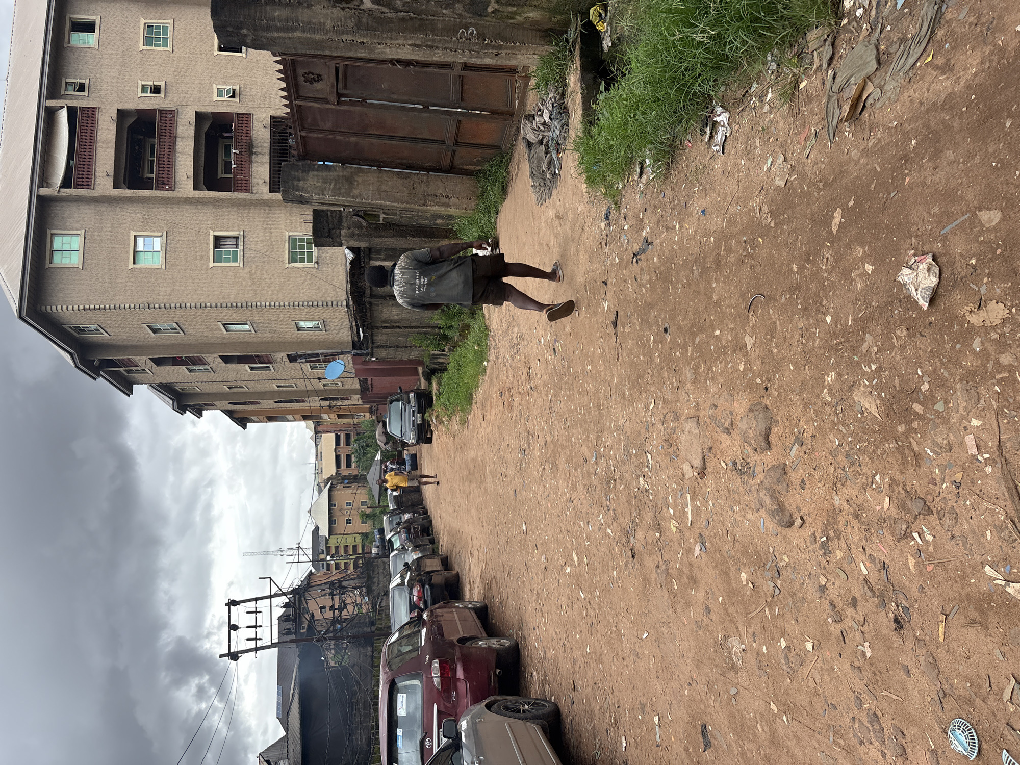 The image depicts a street scene in Nkpor, Nigeria, characterized by a dirt road and overcast skies. In the foreground, a man walks away from the camera, wearing a dark t-shirt with white text and brown shorts. The road is lined with parked cars on the left side of the image. Further down the street, another man in a yellow shirt walks towards the parked cars. Buildings, including a multi-story apartment complex with balconies and several smaller structures, line the right side of the road. The environment shows signs of wear and tear, with debris scattered along the roadside and on the ground. A large, rusted metal gate is visible on the right, adjacent to a concrete wall. The overall impression is one of a densely populated urban area with limited infrastructure.