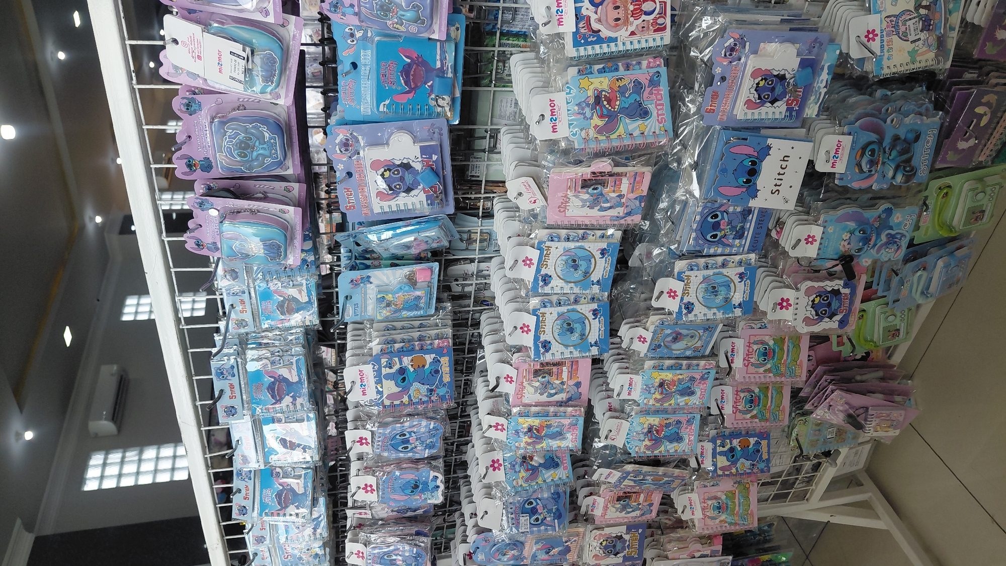 The image depicts a retail display of merchandise featuring the character Stitch from Disney's Lilo & Stitch. The items, primarily notebooks and stationery, are packaged in clear plastic and hung on wire racks. The display is organized by product type and character theme. The background shows interior elements of a store, including fluorescent lighting, a white ceiling with recessed lighting, and a ventilation unit. The overall impression is that of a well-stocked retail environment selling character-themed merchandise, likely in a store located in Purwokerto, Indonesia, given the context.