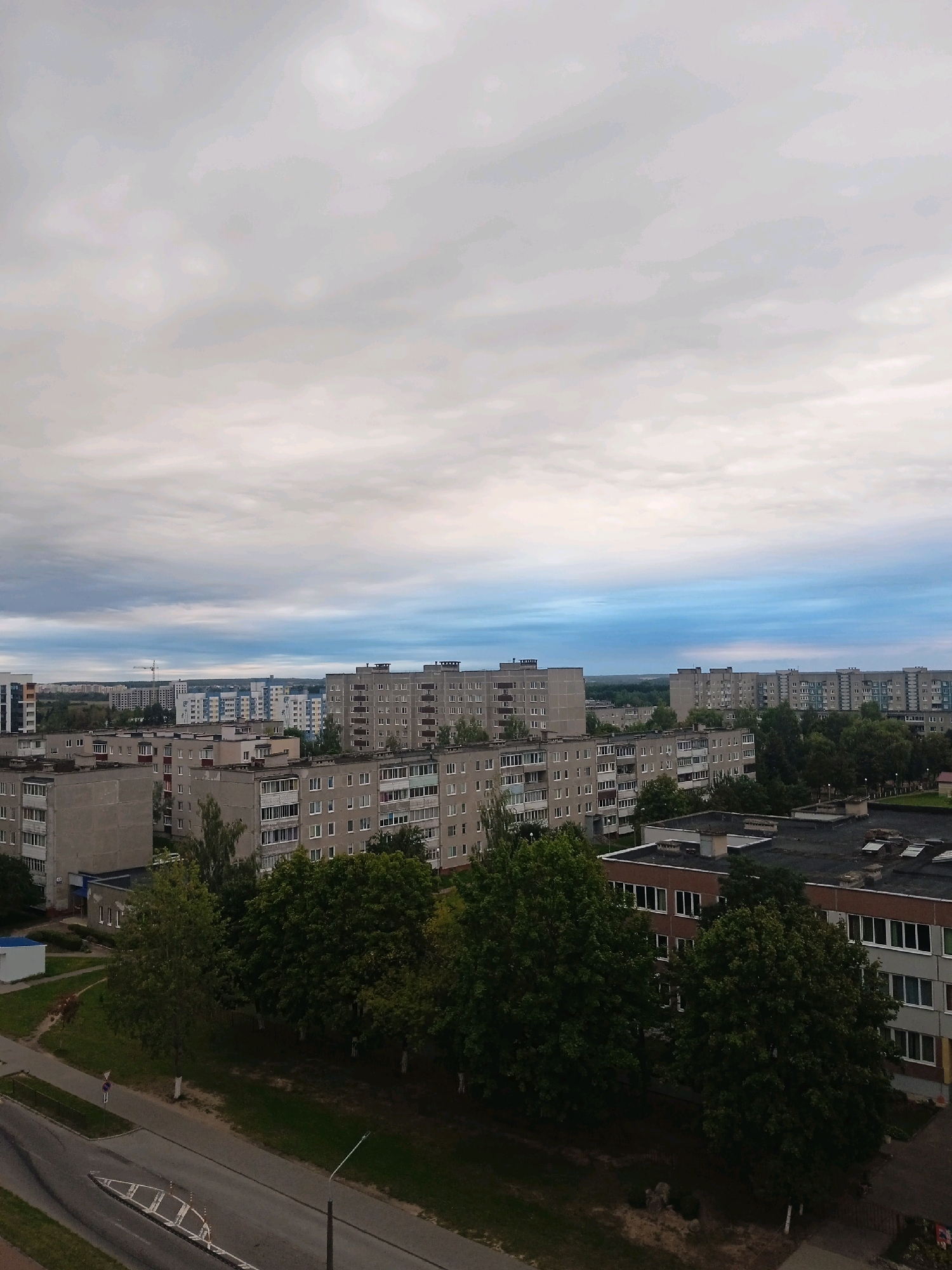 The image shows an aerial view of a residential area in the city of Lida, Belarus. The scene is dominated by numerous Soviet-era apartment blocks, characterized by their grey concrete facades and repetitive window patterns. Several of these buildings are multi-story, with balconies visible on many of them.
In the foreground and mid-ground, trees with green foliage are interspersed among the buildings. Some of these trees are quite mature and large, casting shadows on the ground. A road or street runs through the lower part of the image, with what appears to be a sidewalk or paved area adjacent to it. A lamppost stands on the right side of the road.
In the background, more apartment buildings are visible, extending towards the horizon. A construction crane is also present in the distance, suggesting ongoing development. The sky is overcast with a mix of grey and light blue clouds, indicating a cloudy or potentially rainy day. A few details of human activity are subtly present, such as what might be a person walking on the sidewalk in the lower left quadrant, and a small, parked car or vehicle further in the distance. The overall impression is one of a typical urban landscape in Eastern Europe.
