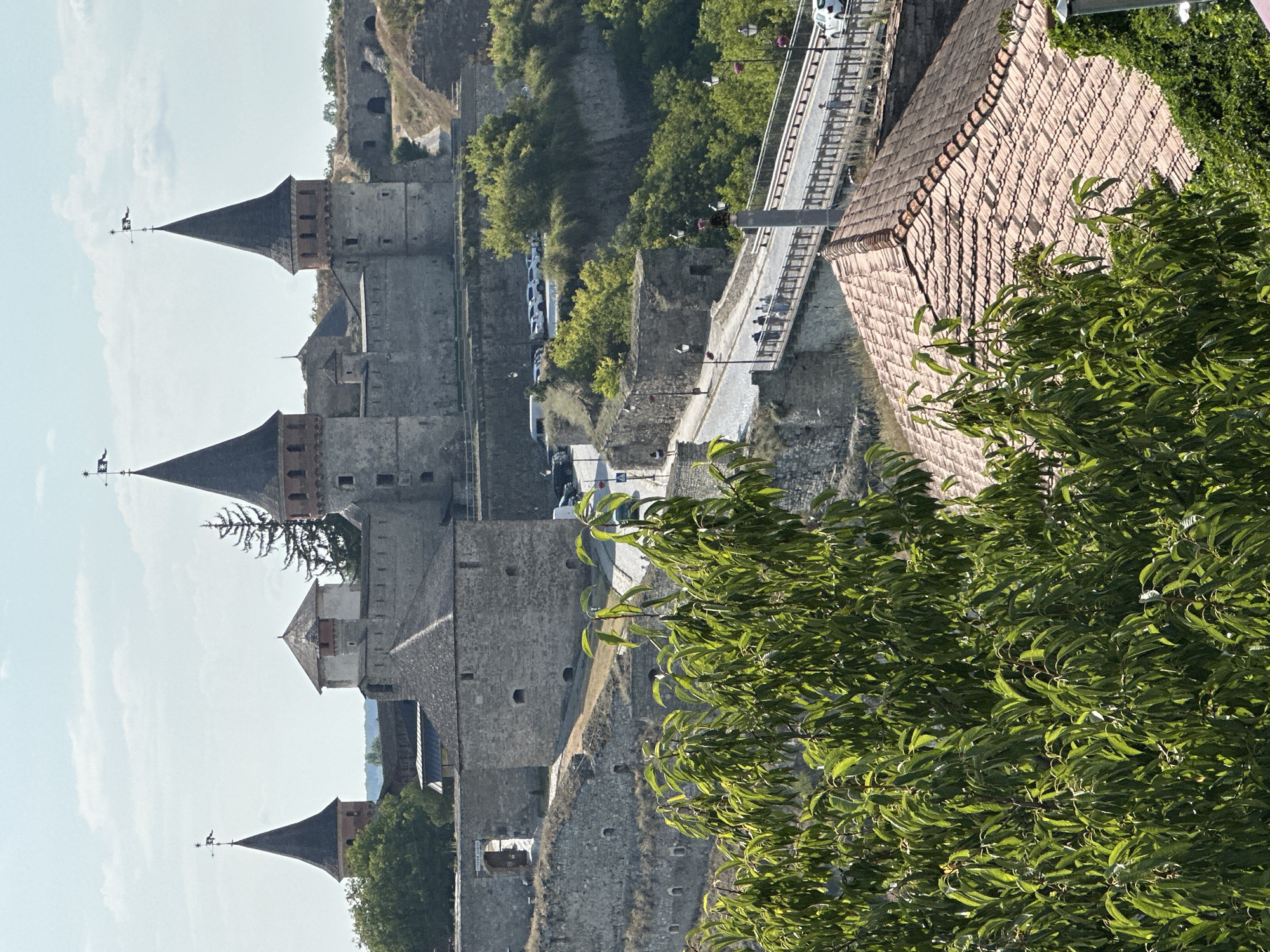 This is a landscape photograph taken from a high vantage point, looking down and across a fortified castle complex in Kamianets-Podilskyi, Ukraine. In the foreground, lush green foliage dominates the lower half of the image, with branches and leaves obscuring parts of the view. Behind the foliage, a terracotta-tiled roof of a building is visible.
The main subject of the photograph is the Kamianets-Podilskyi Castle, a historic fortress built on a rocky island surrounded by the Smotrych River. The castle features several prominent towers with pointed, dark roofs topped with flags. The stone walls and ramparts of the castle are weathered and appear ancient. A road winds its way up to the castle, with several parked cars visible near the entrance.
In the middle ground, a bridge spans a gorge, connecting to the castle area. Several people are seen walking across the bridge. The bridge itself has a metal railing and lampposts. Beyond the bridge, more of the castle's defenses and surrounding terrain are visible, characterized by steep, grassy slopes and rocky outcrops.
The sky is a pale blue with scattered white clouds. The overall lighting suggests a sunny day. The scene captures the grandeur and historical significance of the castle complex.