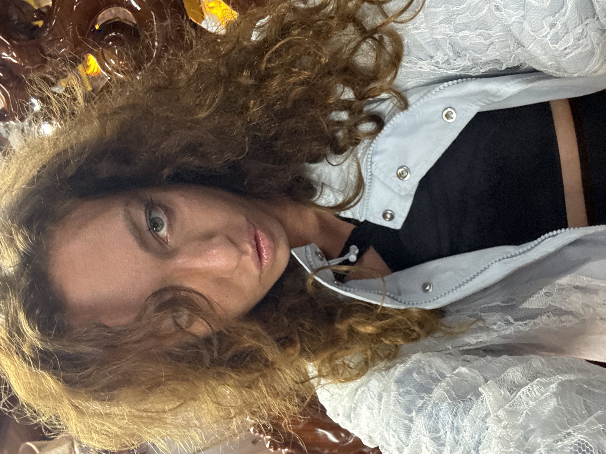A medium shot, selfie-style photo shows a woman with long, curly brown hair. She is wearing a black top and a light blue jacket with lace detailing on the sleeves. Her eyes are green and she is looking directly at the camera with her lips slightly parted. The background appears to be an indoor setting with ornate, carved wooden furniture. The lighting suggests it is either daytime with artificial light or nighttime. There is no indication of the specific location within Mộc Châu, Vietnam.