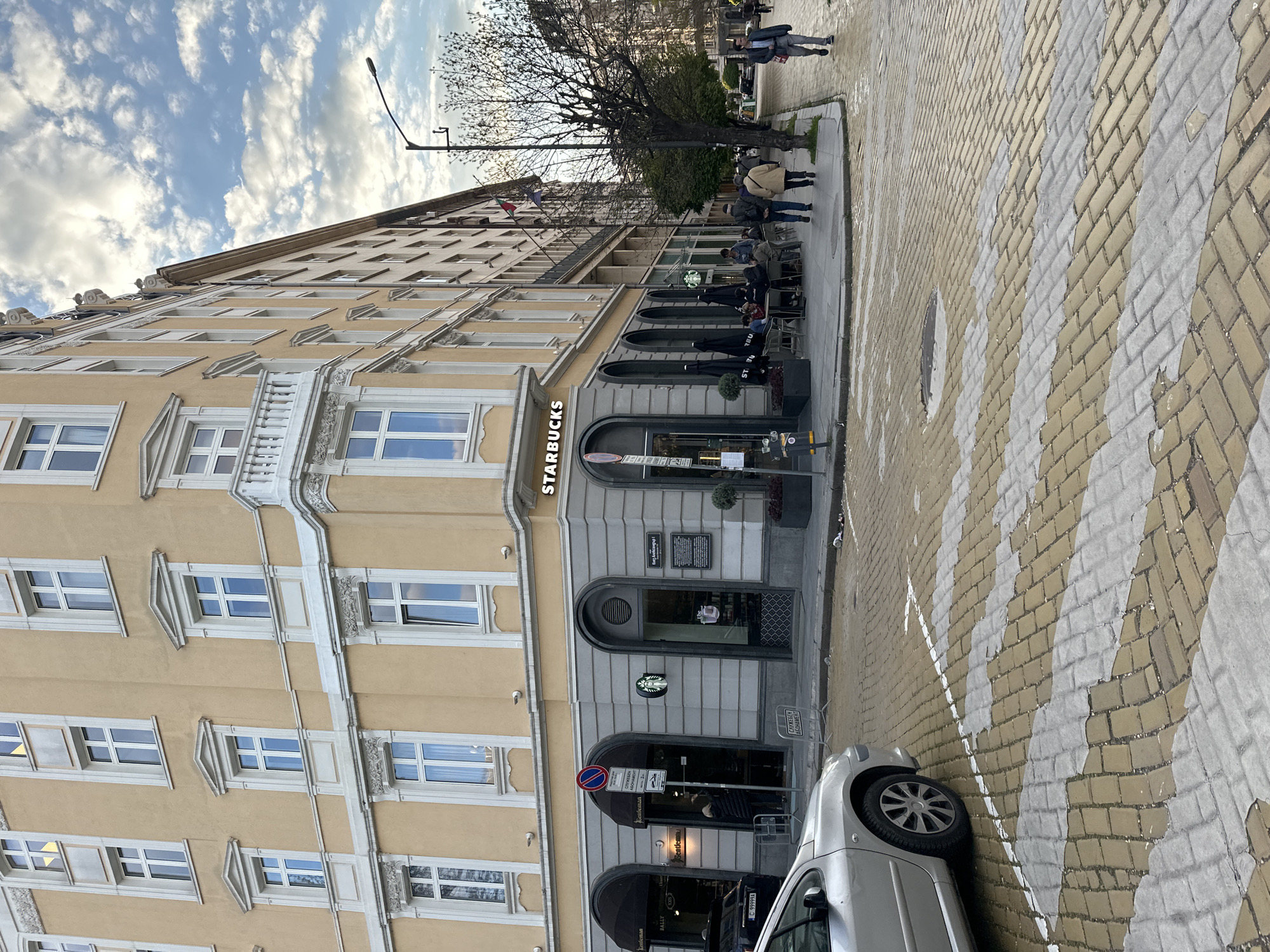 This image captures a vibrant street scene in Sofia, Bulgaria, featuring a multi-story building with a prominent Starbucks coffee shop on its ground floor. The building, constructed in a classic European architectural style, boasts a light beige facade adorned with decorative white window frames, balconies, and cornices.
On the street level, the Starbucks store is visible with its characteristic green logo and lettering, "STARBUCKS." An outdoor seating area extends from the cafe, furnished with black tables and umbrellas, under which several patrons are seated, enjoying beverages. Adjacent to Starbucks, another establishment is indicated by signs bearing the text "Gentleman" and "BALLY."
Several people are seen on the paved sidewalk; some are seated at the cafe, while others are walking, contributing to the everyday urban ambiance. A large, deciduous tree with bare branches stands further down the street, indicating a season other than peak summer, possibly late autumn or early spring. A silver car with license plate "C 99999A" is parked near the curb, partially on the patterned pavement.
The street and sidewalk are distinctively paved with a mosaic of light yellow and white interlocking stones, suggesting a designated crosswalk or a decorative pattern. The sky above is partly cloudy, allowing bright daylight to illuminate the scene, casting soft shadows.
Visible text includes "STARBUCKS" prominently, "Вяз Александър 1" on a plaque on the building (likely denoting "Vaz Alexandar 1"), and "СЛУЖЕБЕН АБОНАМЕНТ" (meaning "Service Subscription" or "Reserved Parking") on street signs. Partial text like "STI BU" and "RBU" is also visible on the Starbucks umbrellas. The overall impression is one of a typical, active urban environment.