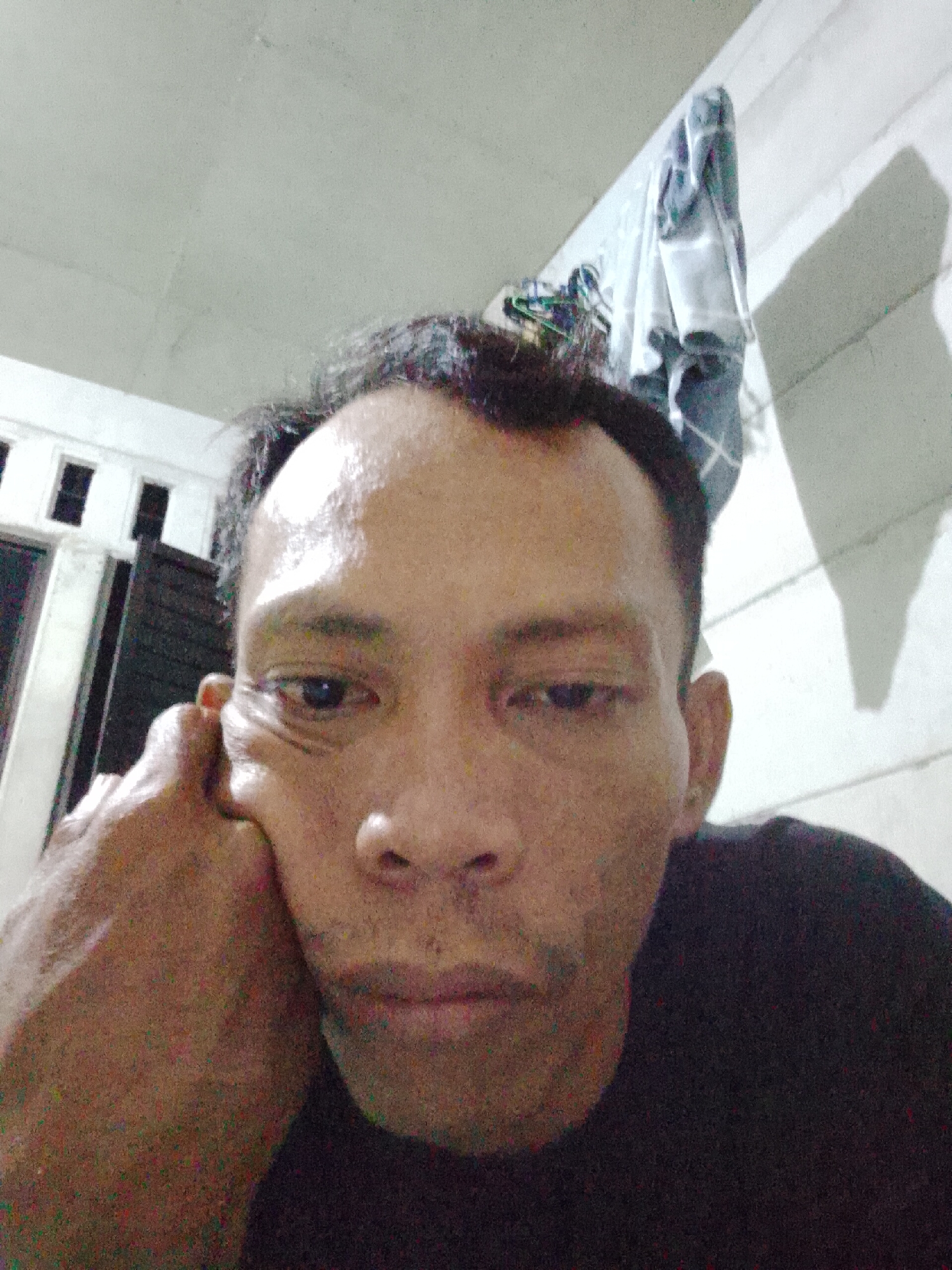 A close-up, indoor selfie shows an adult male with dark hair, resting his head on his hand. His complexion is tan. He is wearing a dark-colored shirt. Behind him are white walls. A window is visible in the background. Clothing hangs on the wall. The picture was taken in Bukit Ketok, Indonesia.