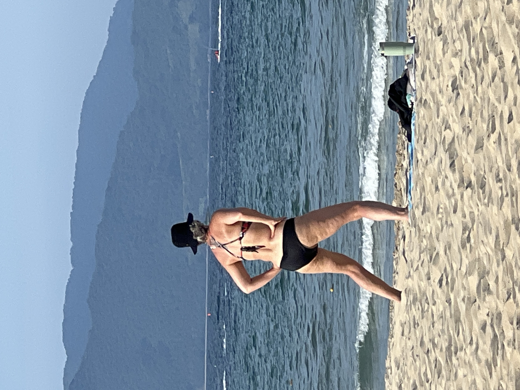 A woman wearing a black hat and a black and floral bikini stands on a sandy beach with her back to the camera, facing the ocean. Her hands are placed on her hips. The ocean is calm with gentle waves rolling onto the shore. In the distance, a speedboat is visible on the water. Behind the ocean, dark blue mountains form the horizon under a clear, light blue sky. On the sand near the woman, there is a blue towel, a black garment, and a green water bottle. The scene appears to be a serene beach setting, likely in Đà Nẵng, Vietnam, given the presence of mountains in the background and the typical coastal environment.