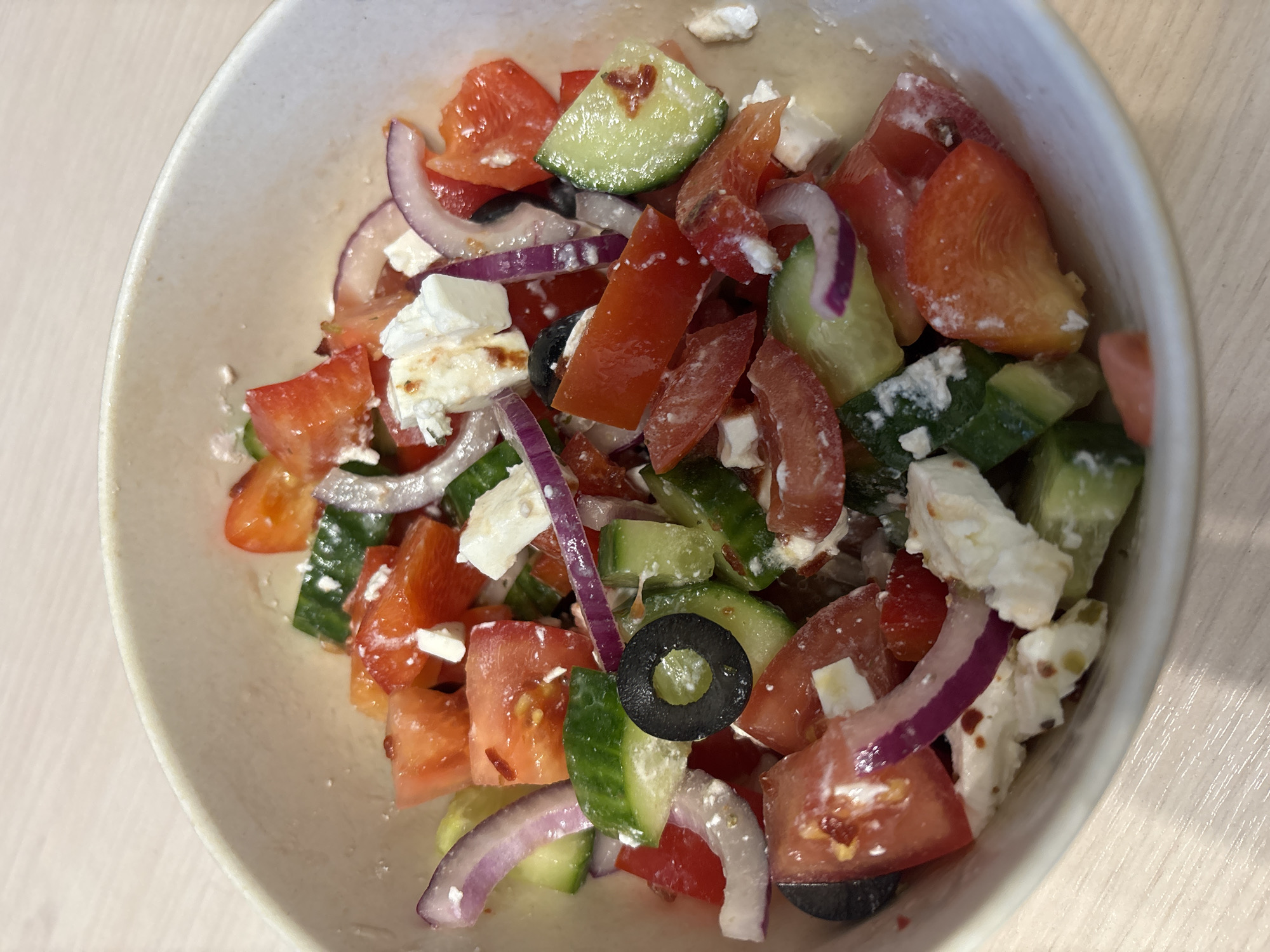 A close-up shot shows a bowl filled with Greek salad. The salad contains diced tomatoes, sliced cucumbers, red onion rings, black olives, and crumbled feta cheese. The bowl is light beige, and the background is a light-colored surface. The image focuses solely on the bowl of salad, and there are no people or indications of the city of Fedorino, Russia.
