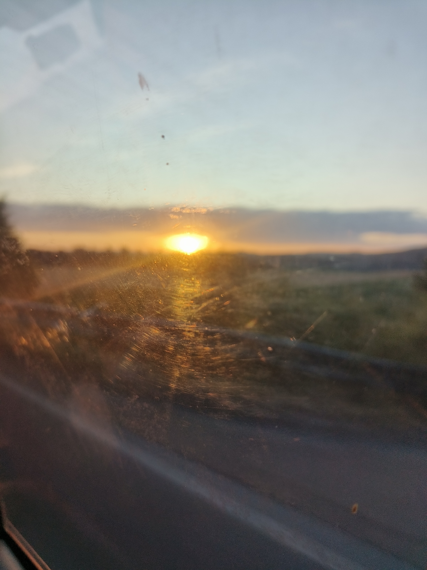 The image depicts a hazy sunset viewed through a dirty window, likely from inside a vehicle. The sun, a bright orange orb, is partially obscured by clouds and casts a warm glow across the landscape. The foreground is dominated by the reflection and smudges on the windowpane, which distort the view of the distant, blurred scenery. The scene is devoid of any people or explicit city structures, suggesting a rural or semi-rural setting on the outskirts of Radobyčice, Czechia. The overall impression is one of a fleeting, melancholic moment captured during twilight.