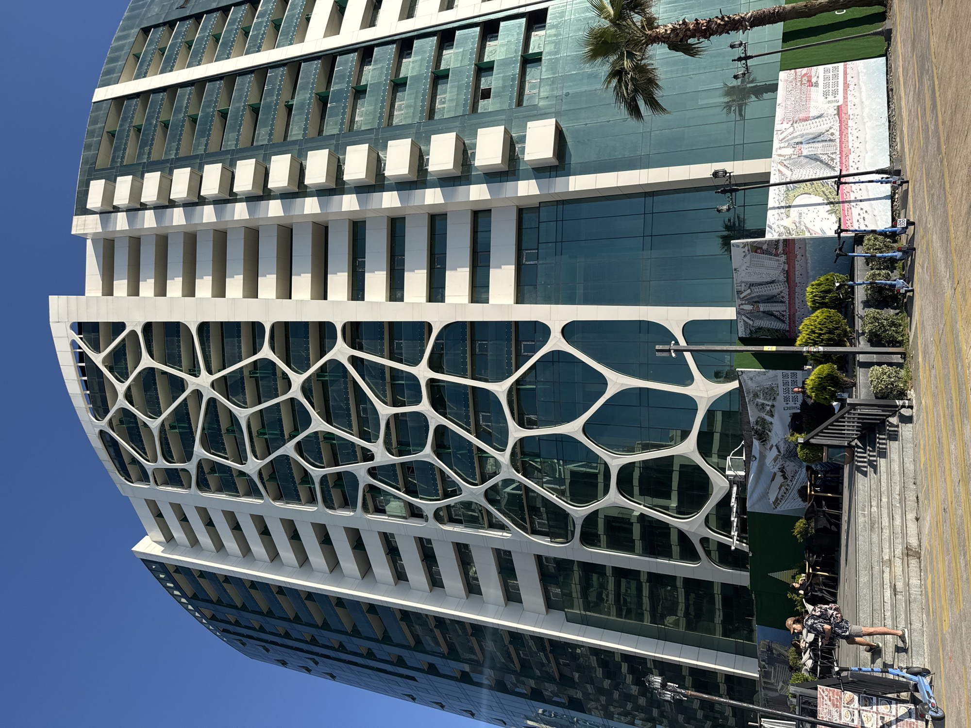 The image depicts a tall, modern building in Batumi, Georgia, showcasing distinctive architectural features. The building's facade is characterized by a white, honeycomb-like structure over glass panels, with alternating sections of regular windows and protruding white elements along the upper floors. A palm tree and advertising signage are present adjacent to the building. A set of stairs leads to a sidewalk area where a few people are standing and walking. Several parked blue scooters are lined up along the sidewalk. The sky is a clear blue, indicating a sunny day.