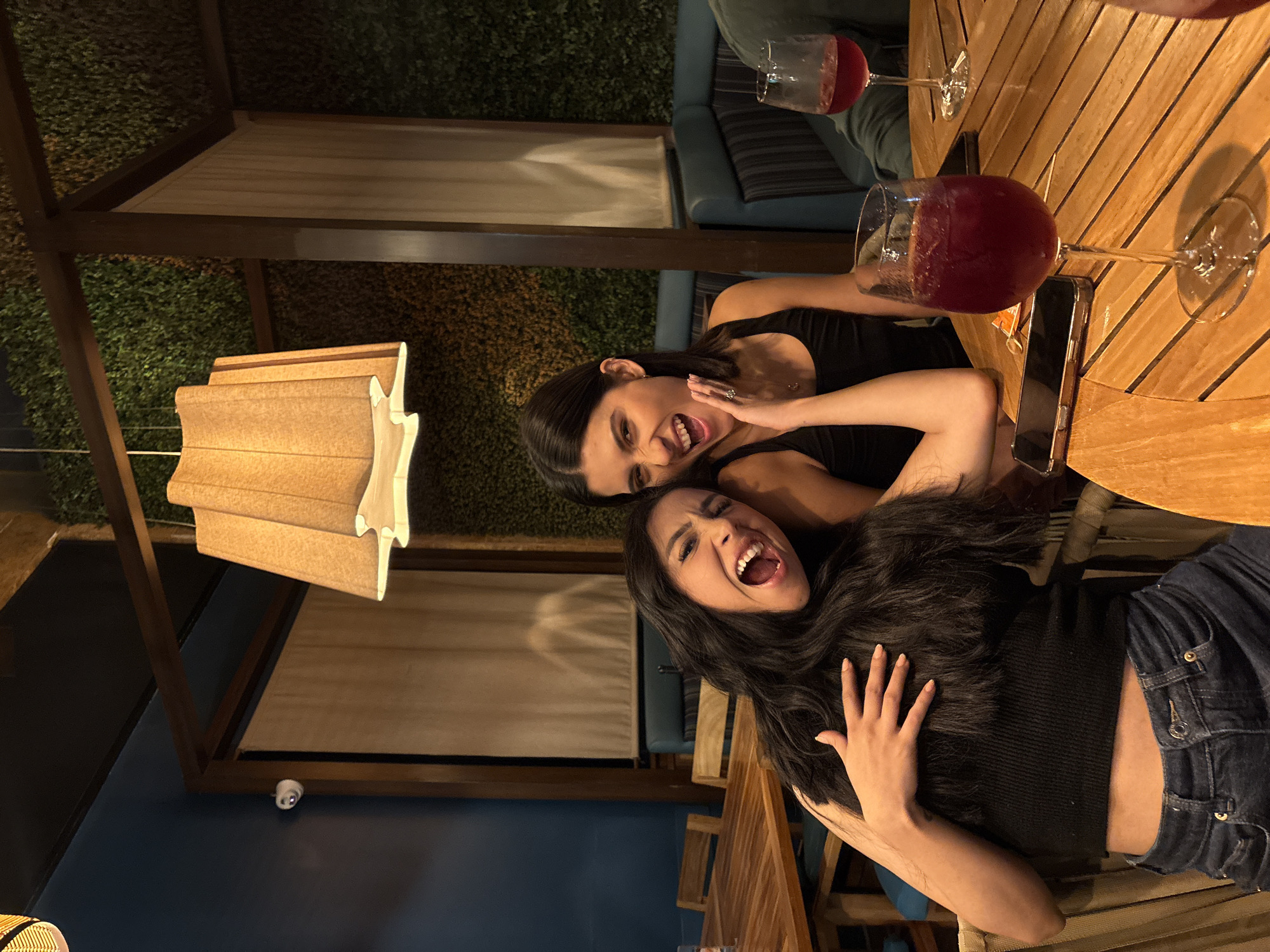 Two young women are at an outdoor restaurant or bar, seated at a wooden table with two large glasses of red liquid, possibly wine or sangria. One woman is wearing a black top and blue jeans, with a visible stomach and her hand resting on her torso. She has long, dark hair and is smiling widely with her mouth open. The other woman is wearing a black sleeveless top and is positioned behind the first woman, with a slightly angry expression. A smartphone is on the table. The background consists of a wall covered in greenery, a light-colored draped awning, and wooden framework, suggesting a covered patio or outdoor dining area. A modern, stylized lamp hangs from the ceiling. The city of Puebla, Mexico is not discernable from the image provided.