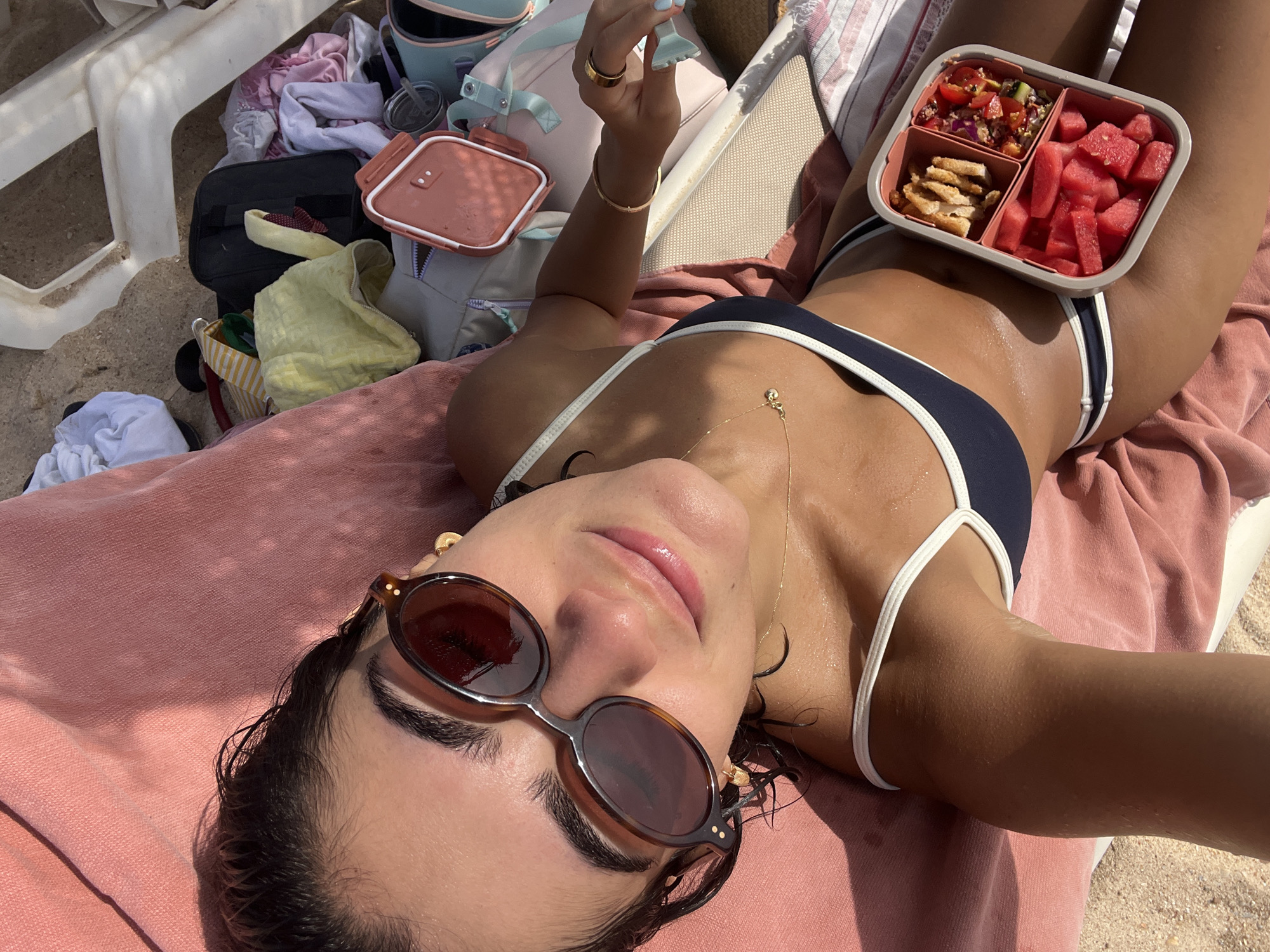 A young woman with dark hair and sunglasses is lying on a pink towel on a beach chair. She is wearing a dark blue and white bikini. In front of her, to her right, is a bento-style lunchbox containing cubed watermelon, a salad, and some croutons. Several items of clothing and bags are scattered around her on the sand and chair. The scene appears to be outdoors, likely at a beach location near Almancil, Portugal, given the bright sunlight and beach setting.