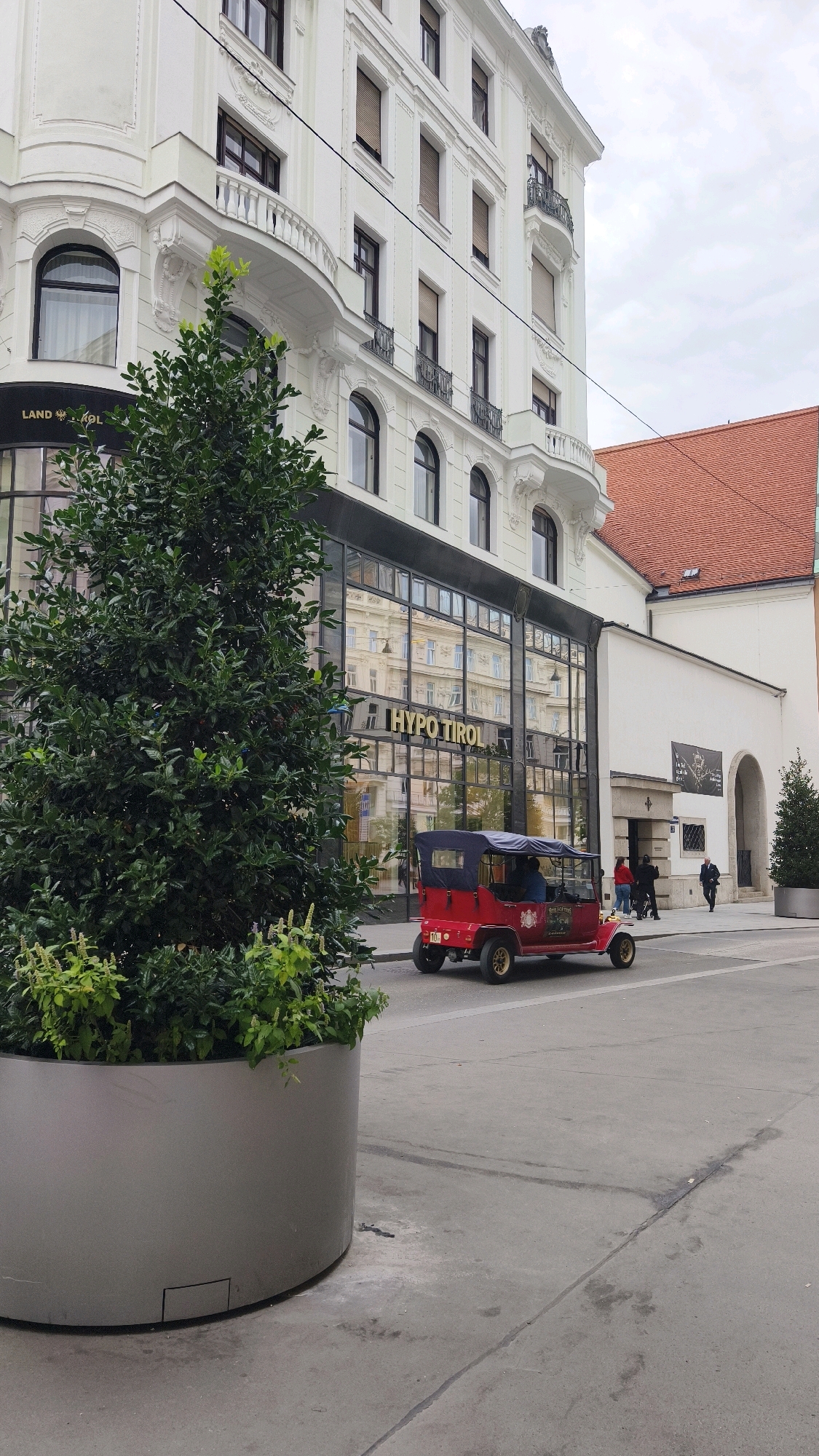 An outdoor, eye-level, medium shot captures a street scene in Vienna, Austria. In the foreground to the left, a large, metallic grey planter holds a dense, green shrub. Behind it, the facade of a white, multi-story building dominates the frame. The building features classical architectural elements, including ornate balconies with wrought iron railings and arched windows with dark frames. The ground floor has a modern glass facade with the text "HYPO TIROL" prominently displayed in gold lettering.
On the street in front of the building, a red, vintage-style convertible car is parked. It has a dark blue canopy and two visible occupants. A few people are walking on the sidewalk in the midground, some approaching the entrance of the building, which has a large arched doorway. To the far right of the image, a smaller portion of a building with a red tiled roof is visible, and a portion of a Christmas tree is visible. The sky is overcast and grey.