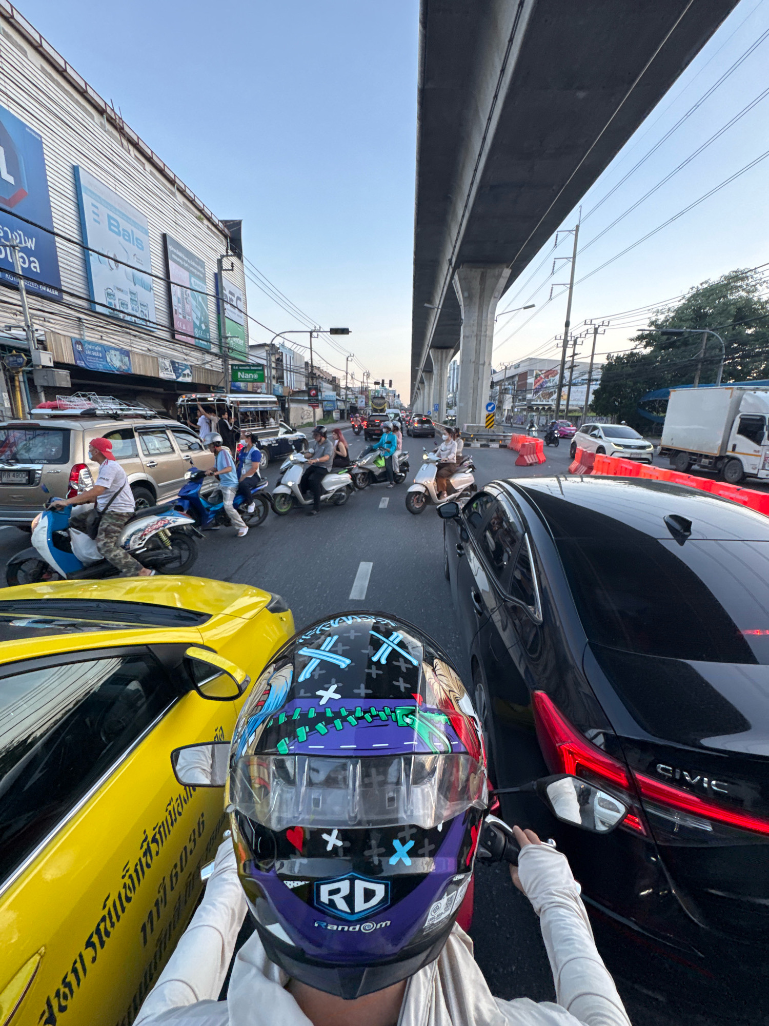 The image depicts a traffic scene in Samrong Nuea Subdistrict Municipality, Thailand, under an elevated highway structure. In the foreground, a person wearing a motorcycle helmet with a decorative design and light-colored arm sleeves is seated on a vehicle, holding a mirror. To the left, a yellow car is visible, with Thai text on its side. To the right, a black sedan with "CIVIC" branding is in the frame. The road is occupied by various vehicles, including scooters with riders, a tan SUV, a bus, and a white truck. Signs and buildings line the street, and overhead power lines are present. The sky is clear and light blue, suggesting it is either early morning or late afternoon. The overall scene indicates congested urban traffic.