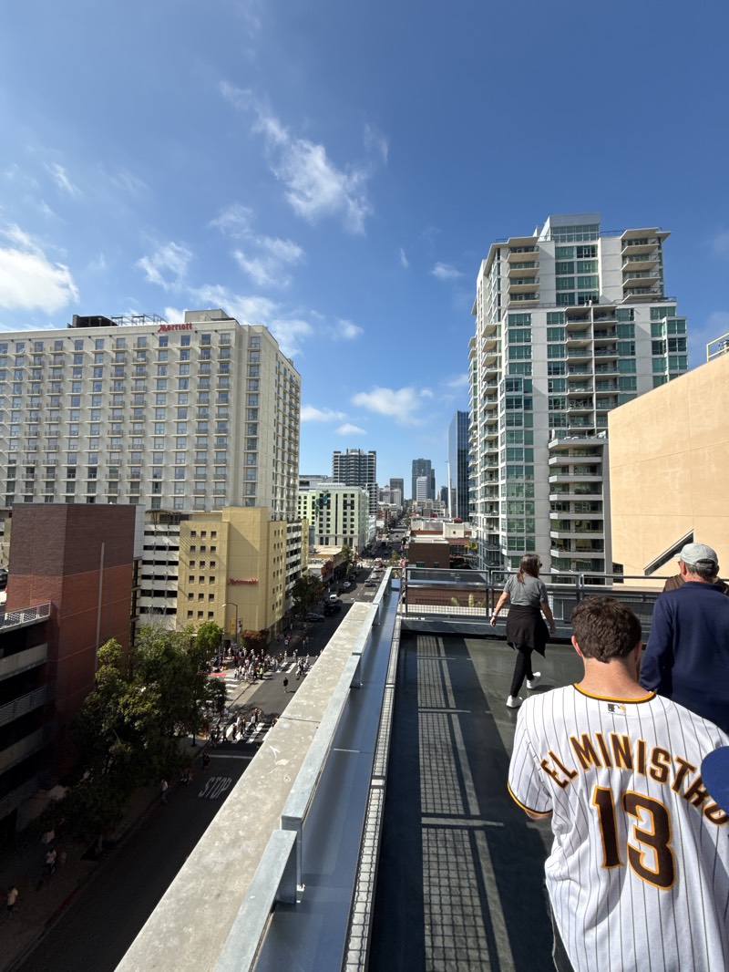 The image depicts a view from a rooftop in San Diego, United States. The foreground contains two people on the rooftop. The person closest to the camera is wearing a baseball jersey with the number "13" and the name "EL MINISTRO" on the back. Further down the roof, a woman in a grey top and black jacket walks in the same direction as the other two figures.
The backdrop features a cityscape of San Diego under a blue sky with scattered clouds. Tall buildings dominate the view, including a large Marriott hotel on the left and a modern apartment or office tower on the right. A street scene is visible below, with pedestrians crossing a crosswalk and moving along the sidewalks. The buildings in the background extend into the city center.