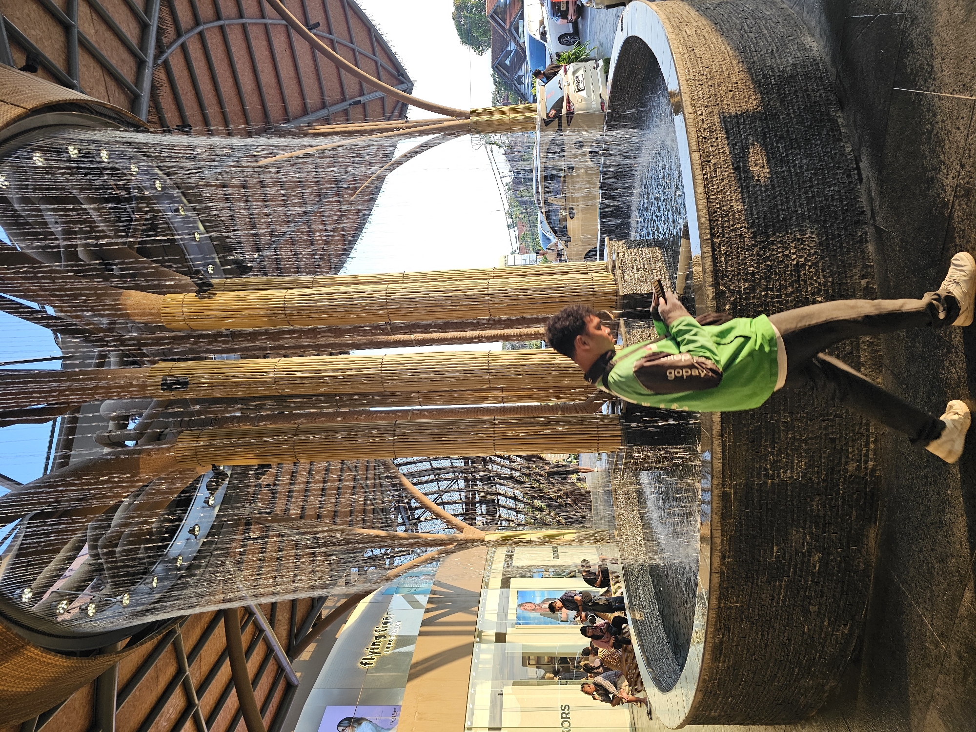 In Denpasar, Indonesia, a man in a green hoodie and dark pants walks in front of a circular fountain with water cascading from above. The fountain is surrounded by a dark stone wall. Behind the fountain, large bamboo-like structures support the cascading water, which falls from a curved, woven ceiling. In the background, people are seated on a low wall near shop fronts, including one with a "Flying Tiger Copenhagen" sign. Cars are parked in an outdoor area beyond the shops, and residential buildings are visible in the distance. The lighting suggests it is either early morning or late afternoon.