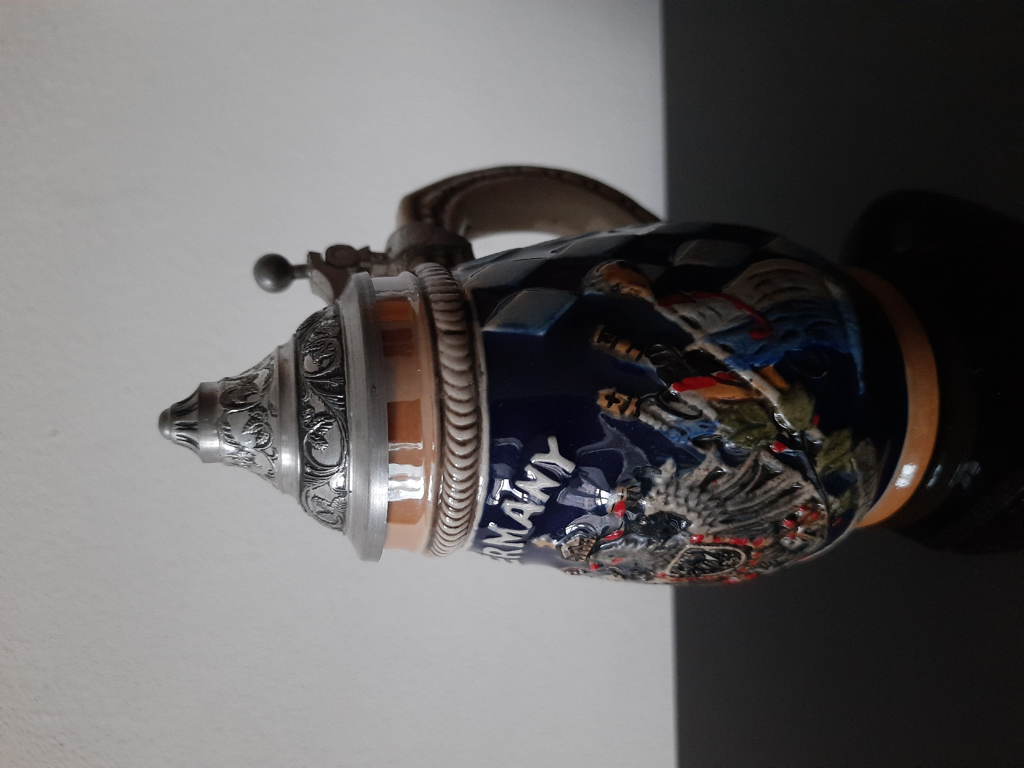 A ceramic beer stein with a pewter lid sits on a black reflective surface in front of a plain white wall. The stein is decorated with a dark blue base, a lighter brown band below the lid, and relief decorations of figures in historical costumes. The word "GERMANY" is visible in white lettering on the blue portion of the stein. The lid is ornate with embossed patterns and a small finial. A handle is attached to the side of the stein. The background is a simple, unadorned wall. The lighting is soft and diffused, casting a slight shadow from the stein onto the surface. The image does not contain any elements that suggest a specific location in Lviv, Ukraine.