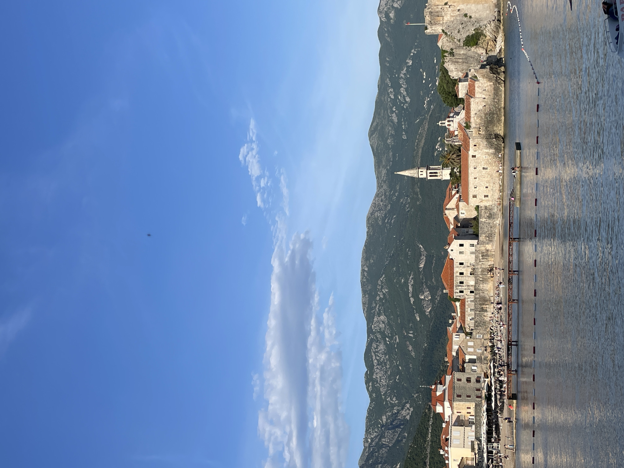 The image depicts a view of the coastline of Budva, Montenegro. The sky is mostly clear with some thin, elongated clouds. The mountainous terrain rises steeply behind the buildings along the shore. The buildings feature light-colored walls and red-tiled roofs, with a tall church tower among them. A pier extends into the water, marked with alternating red and white buoys. The water's surface is rippled, and a boat is visible in the top right corner. People are visible on the pier and the beach.