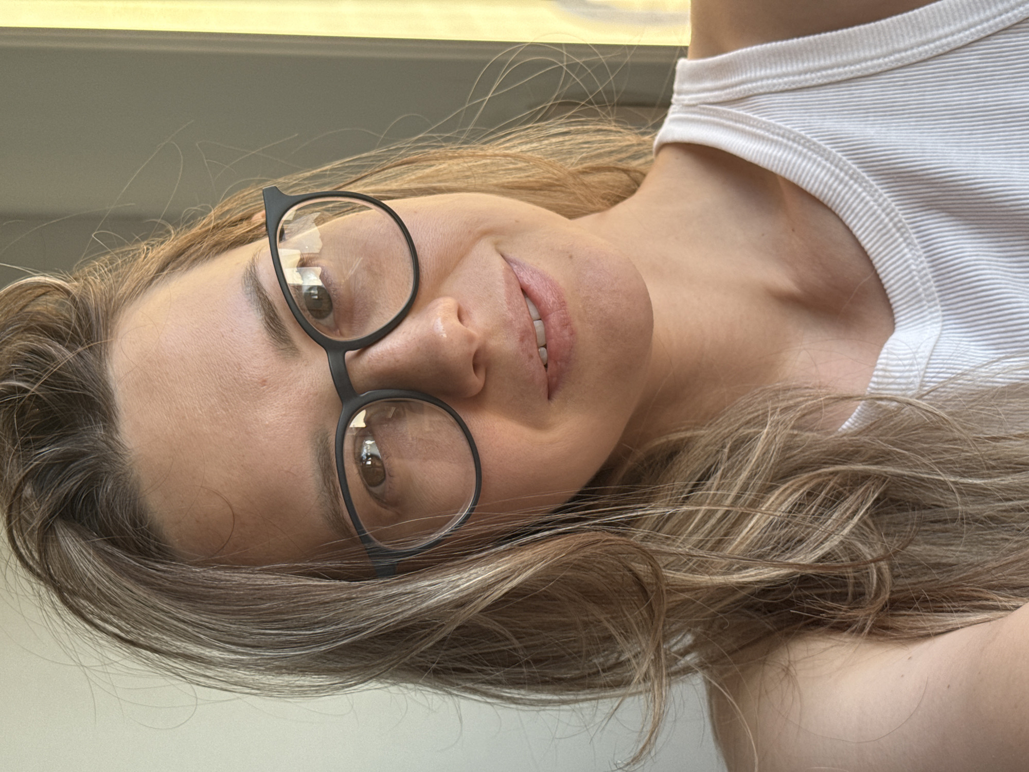A woman with long blonde hair is wearing black framed glasses and a white ribbed tank top. The background is a light, neutral color, possibly a wall or ceiling. The photograph appears to be taken indoors. No information connects the image to the city of Chaikovsky, Russia.