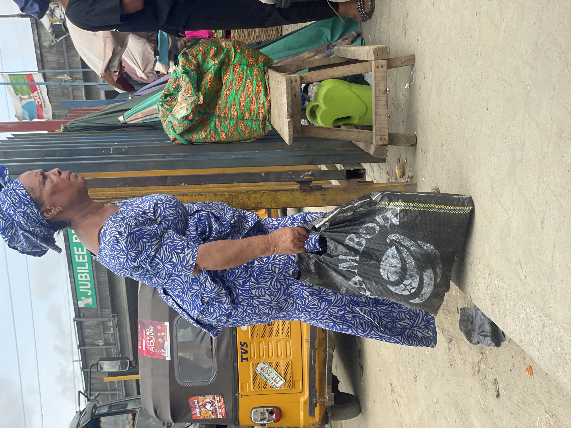 The image captures a lively daytime street scene or outdoor market in Lagos, Nigeria, under an overcast sky. In the foreground, an elderly woman is the central subject, dressed in a distinctive blue and white patterned long-sleeved dress and a matching headwrap. She holds a large black bag featuring white text, "MBOW," and a logo.
Behind the woman, a yellow and black Bajaj RE tricycle, commonly known as a keke napep, is visible. It displays the license plate "LAGOS FST-350P," the brand "TVS," and stickers including "PUNCHLINES" and "Help Above" with "Folarsquare Church" written above it. In the background, there's a wooden stool holding a bright green plastic watering can and a vibrant green patterned fabric bag. The scene is lined with market stalls or temporary structures, featuring various awnings and tarpaulins, suggesting a busy commercial area.
Other individuals are partially visible in the background, reinforcing the bustling atmosphere. A white flag with a green and red logo and the text "OTA" is seen in the upper background. A green sign with white text, "JUBILEE P" (likely part of "JUBILEE PLAZA"), is also visible. The overall setting depicts a typical, active urban environment in Nigeria.