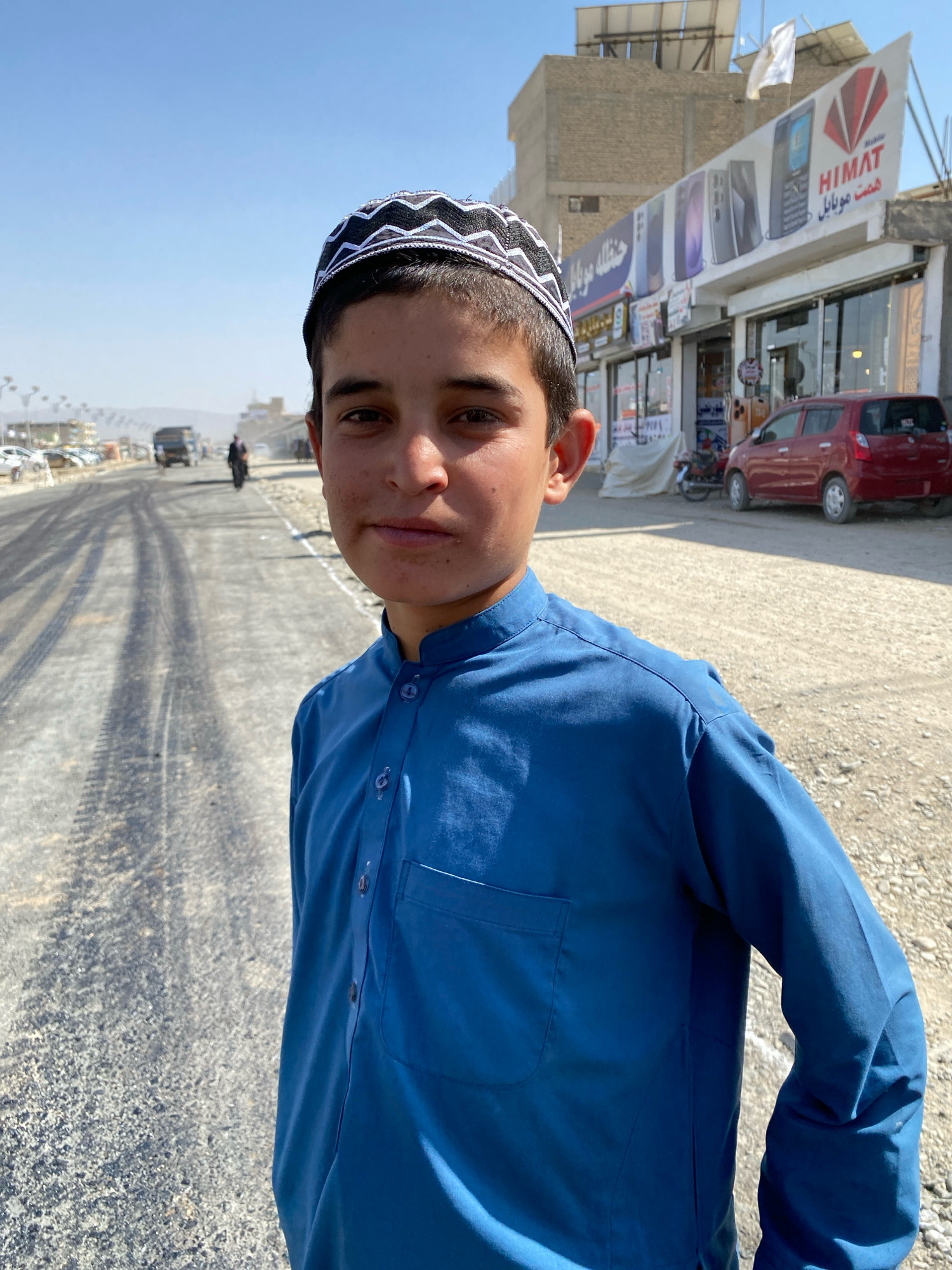 A young boy, wearing a blue shalwar kameez and a white and black patterned kufi, stands on a dusty road. The boy faces the camera with a neutral expression, his gaze directed forward. Behind him, the road stretches into the distance, marked by tire tracks and bordered by a gravelly shoulder. In the background, a few vehicles, including a truck and a red car, are visible. A building with a prominent banner displaying "HIMAT" and Dari text is also present. The scene is set under a clear blue sky, suggesting a bright, sunny day. The overall atmosphere appears to be that of a bustling street in Spin Boldak, Afghanistan, likely during daylight hours.