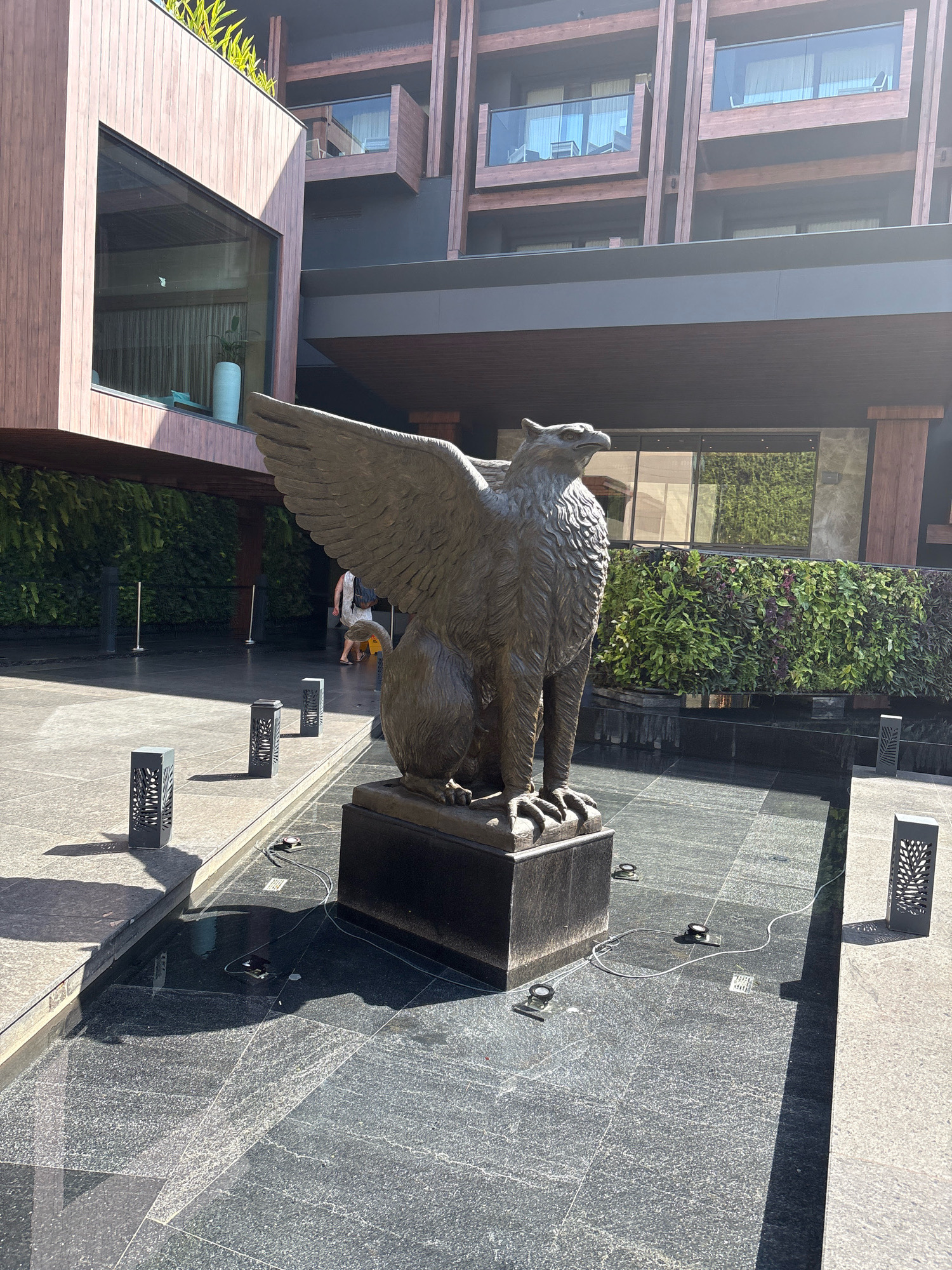 The image depicts an outdoor area with a bronze statue of a griffin as the central subject. The griffin is seated on a dark pedestal, with its wings spread upwards. The statue is located in a courtyard with dark granite-tiled flooring that appears to be a shallow water feature, as evidenced by reflections and ripples. Several dark, rectangular outdoor lights with cut-out patterns are placed around the perimeter of the water feature. In the background, a modern building with wooden and dark gray facade elements is visible, featuring multiple balconies with glass railings. A vertical green wall covered in lush foliage runs along the base of the building. A person wearing a light-colored dress and carrying a dark backpack is walking away from the camera in the middle ground, to the left of the statue. The lighting suggests it is daytime, with sunlight casting shadows on the ground. The overall environment appears to be a resort or hotel, likely in Vagator, India, given the architectural style and lush greenery.