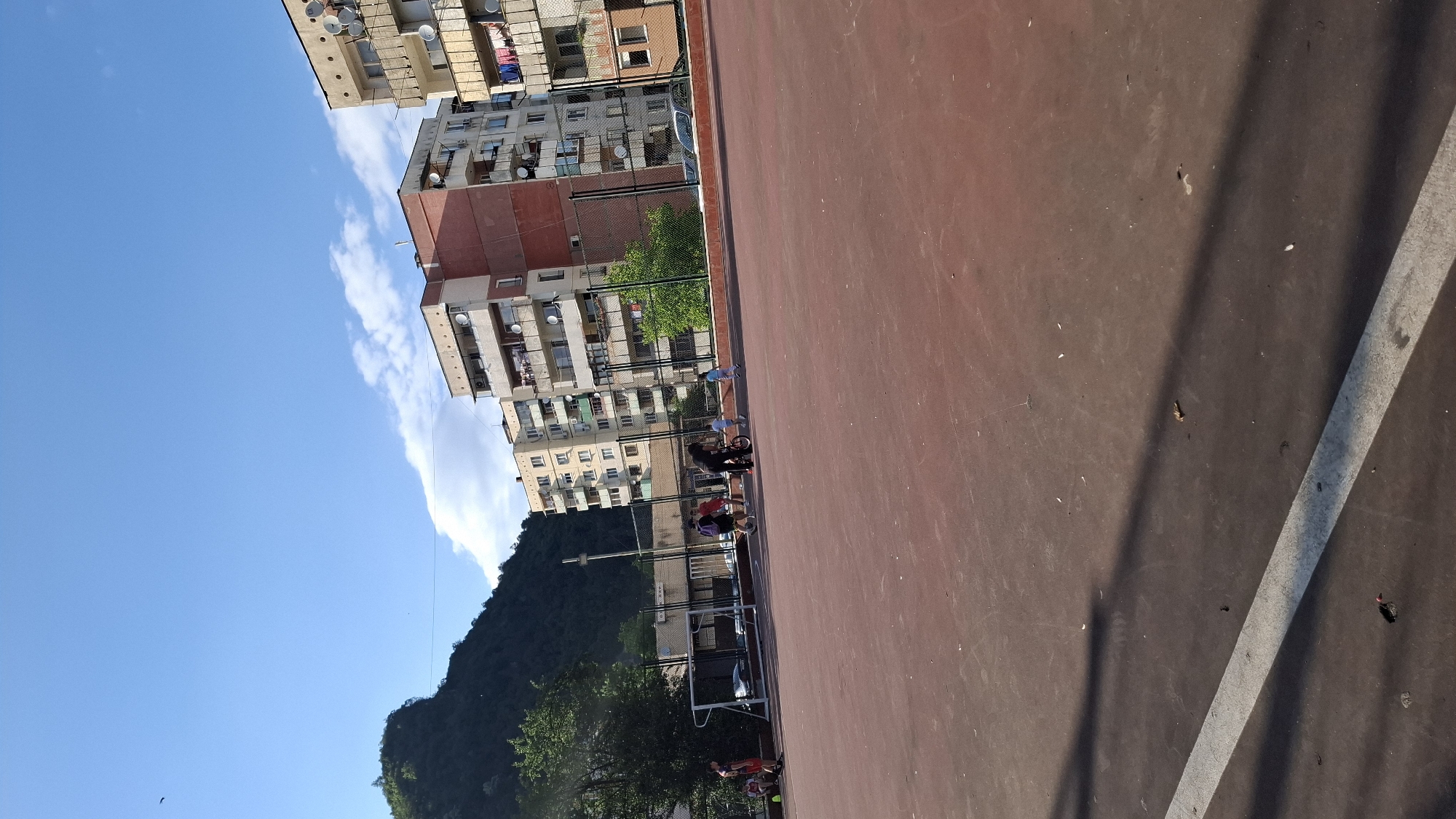 This image depicts an outdoor sports court in an unknown urban or suburban area, at the base of a forested mountain or large hill, under a clear blue sky with scattered white clouds. The time of day appears to be late afternoon or early evening, evidenced by long shadows cast diagonally across the court from the left.
The foreground is dominated by a reddish-brown paved sports court, marked with a single white line running diagonally from the lower left to the middle right. Debris, including small leaves and dirt, is visible on the court surface.
In the midground, approximately seven to eight individuals are present on the court. On the left side, near a white metal goal frame (likely for soccer), two individuals stand; one wears a red shirt and dark shorts, and the other wears a dark shirt and dark shorts. In the center, a person in dark clothing, possibly an adult, pushes a child on a small bicycle. Nearby, another child in a light-colored shirt and dark shorts runs to the right. To the left of the bicyclist, two more individuals walk towards the left; one wears a purple backpack and dark shorts, and the other wears a red shirt. On the far right, a child in a light blue shirt and light shorts runs towards the center. No direct interactions or organized play are clearly discernible, but the individuals appear to be engaged in leisure activities.
Behind the court, a black chain-link fence separates the playing area from a street or parking area. Partially visible beyond the fence are a white car and a dark-colored car. Further back, several multi-story residential apartment buildings of varying designs and colors stand, including one with a reddish-brown section. These buildings feature numerous balconies, many equipped with satellite dishes, and some with laundry hanging. Green trees and bushes are interspersed between the court, fence, and buildings. The background on the left is dominated by a densely forested green hill or mountain, rising significantly above the urban structures. A small dark bird is visible flying in the upper left portion of the sky.
