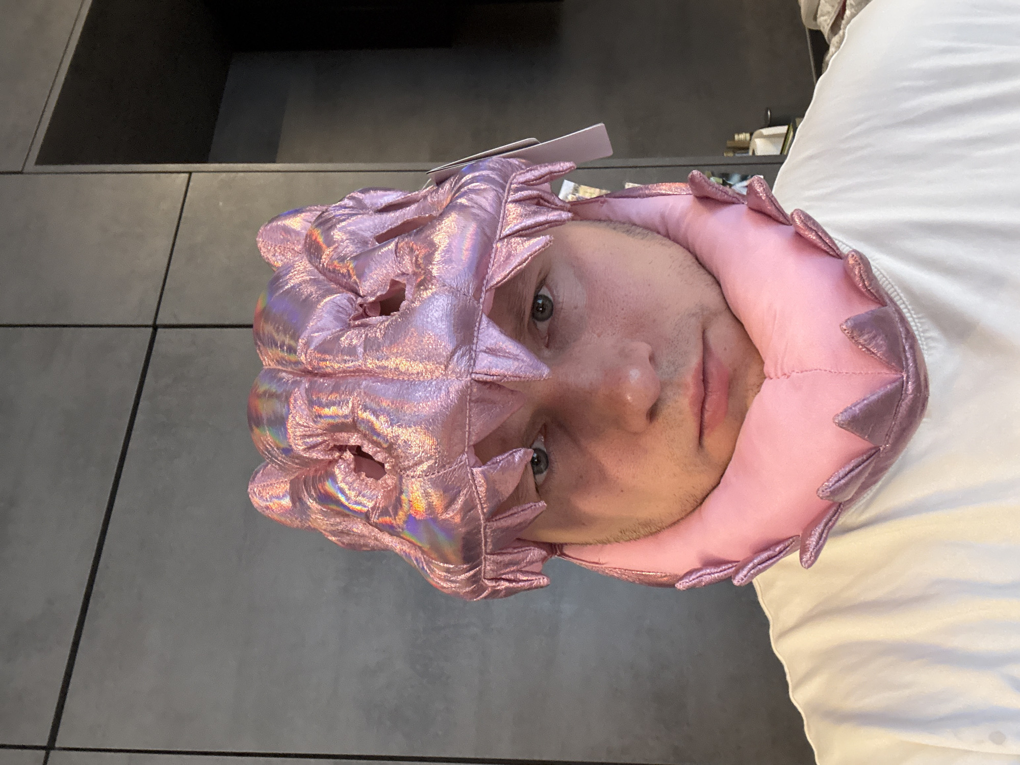 A person is wearing a pink and iridescent head covering. The background includes a grey tiled wall and a white bed. The location is not discernible from the image.