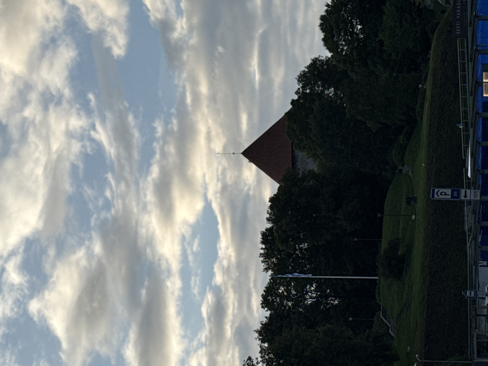 The image shows a view of a prominent conical-roofed tower, likely part of the Old Town of Tallinn, Estonia, rising above a dense line of dark green trees. A flag, appearing to be blue and white, is planted on a flagpole in front of the tower, to the left. The tower is situated on a green, sloped hill with a set of stairs leading up to it. The sky above is a mixture of blue sky and numerous scattered clouds, some appearing bright white and others shaded gray, suggesting it could be late afternoon or early evening. In the foreground, a sign with a blue "P" symbol and "24h" below it indicates parking. Below the sign, a blurred structure of what appears to be a modern building with blue elements is visible. The overall scene depicts a historic landmark within a natural setting, overlooking a more contemporary urban area.