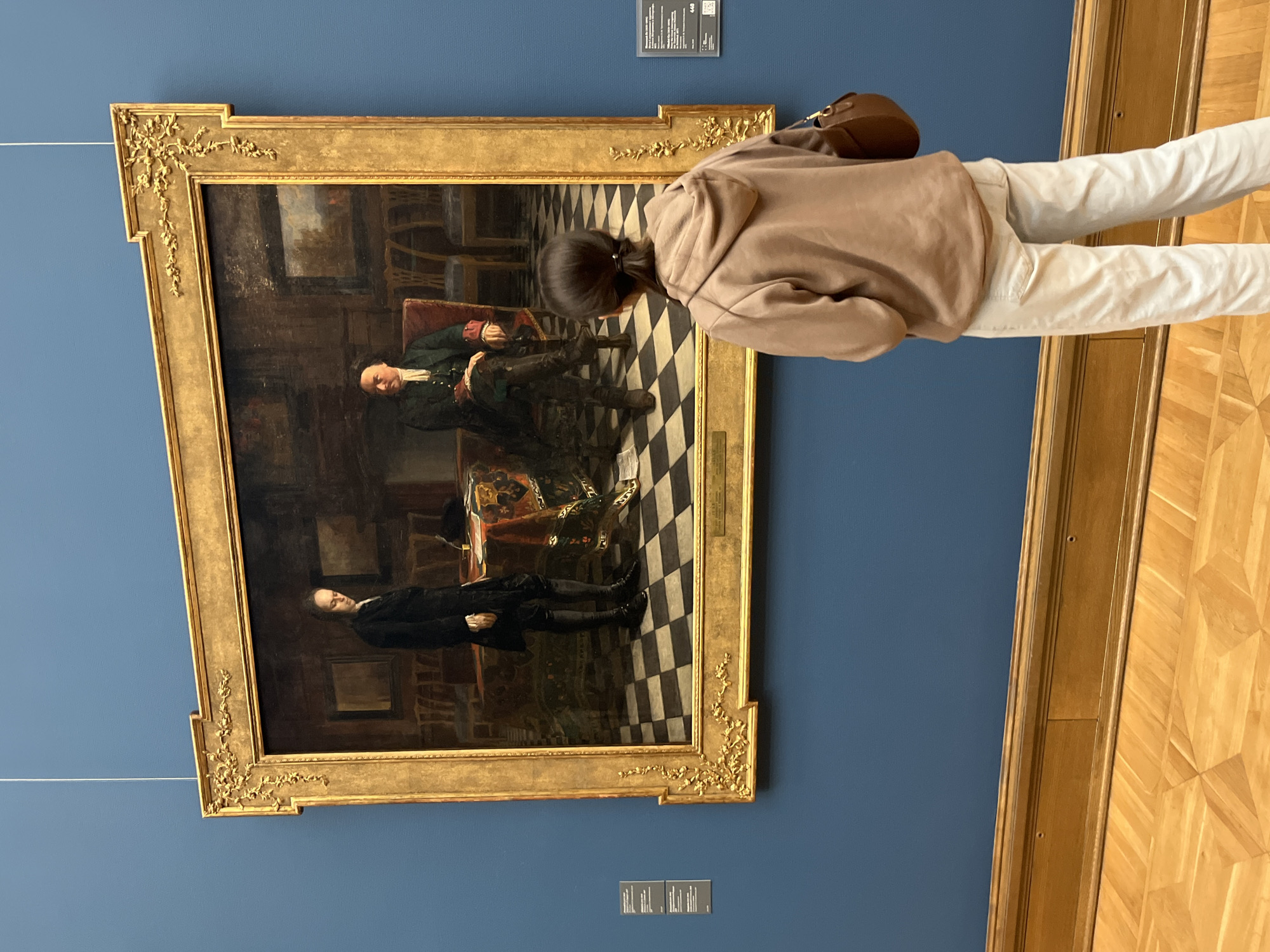 The image shows a young woman with dark hair pulled back in a ponytail, wearing a light brown hooded sweatshirt and off-white pants, standing in front of a large, ornate framed painting in a museum. The painting depicts two men in a formal setting. One man, on the left, stands and is dressed in a dark suit with a white ruffled collar. The other man, on the right, is seated in a chair, wearing a dark green coat with red lapels and a white cravat, and dark breeches. The room in the painting has a checkered floor, a table covered with a patterned cloth, and chairs. The painting is displayed on a deep blue wall, and the floor in the foreground is made of polished wood. To the left of the painting, a small informational plaque is visible. This scene is taking place in the Tretyakov Gallery in Moscow, Russia.