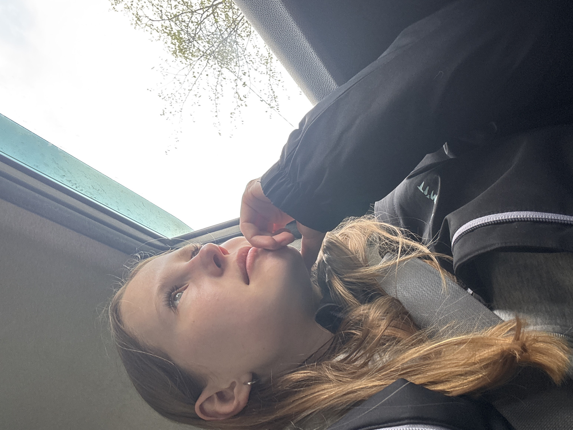 A person is reclining inside a car, looking up through the open sunroof. The person has light skin, blonde hair, and is wearing a black jacket or sweatshirt. Part of the car's interior is visible, including the sunroof and the car's headliner. Through the sunroof, there is a view of a cloudy sky and the branches of a tree with budding leaves. There is no indication of the location being Chaikovsky, Russia.