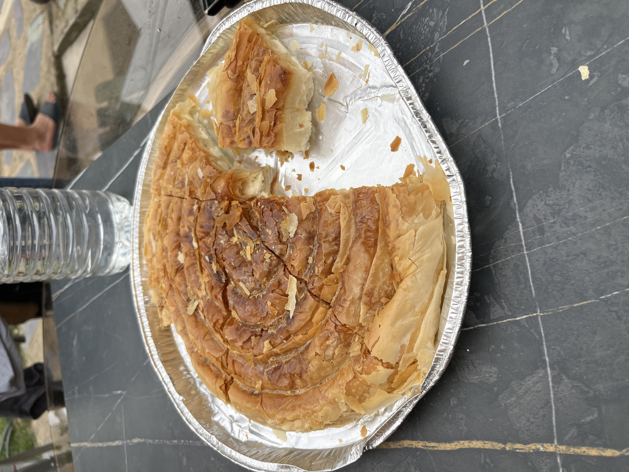 A partially eaten, spiral-shaped pastry with a golden-brown, flaky crust is presented in a round aluminum foil pan on a dark, speckled table. The pastry is sliced into several portions, with one piece removed and placed to the side, revealing a white, creamy filling. Small, light-colored flakes, likely from the crust or topping, are scattered around the pan. Behind the pastry, a clear plastic bottle of water is visible. In the background, out of focus, are the legs of two individuals wearing sandals, standing on a light-colored, textured surface that appears to be an outdoor patio or walkway. The environment suggests an informal outdoor setting, possibly a backyard or terrace, in Stains, France, given the information provided.