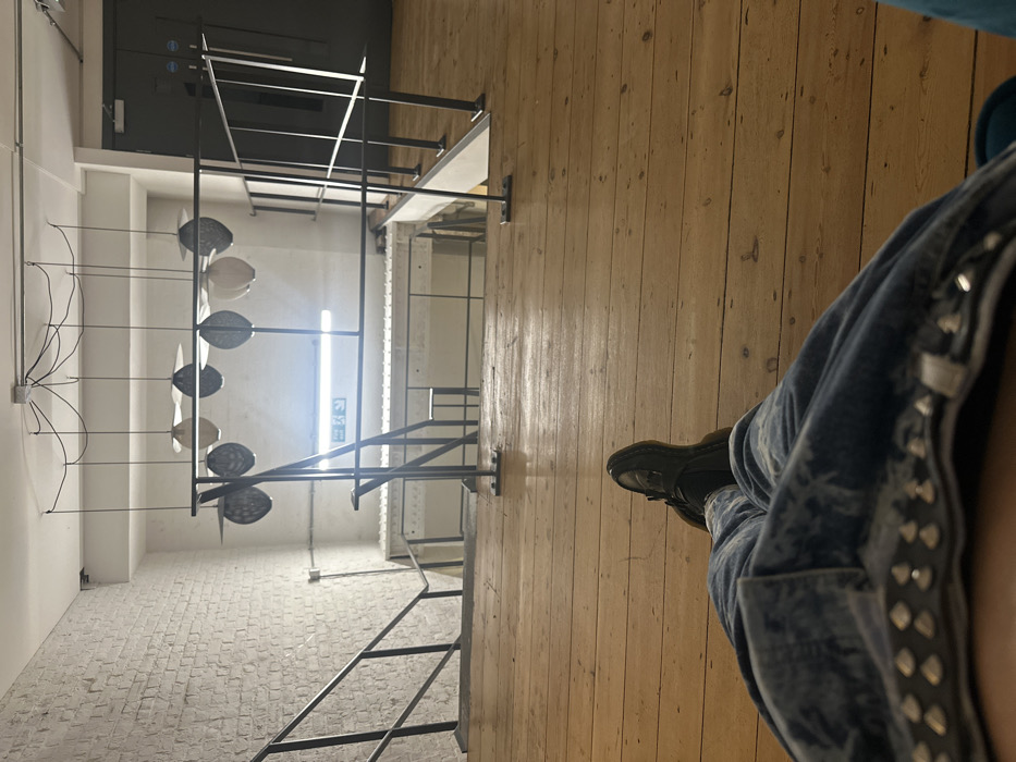 The image displays an interior view from a first-person perspective, with the viewer's lower body visible in the foreground. The individual is wearing distressed blue jeans with a studded trim along the waistband and dark, possibly black, flat shoes or loafers. The legs are extended onto a light-colored wooden floor composed of planks.
The scene looks down into a multi-level contemporary space. On the left, a white-painted brick wall ascends from a lower level, alongside a dark metal staircase with railings that descends out of view. In the mid-ground, a central void reveals a lower floor, bordered by dark metal railings supported by vertical posts. Beneath this railing, a further set of stairs or a similar architectural feature is partially visible.
Above the lower level, suspended from a white ceiling, hangs an art installation comprising approximately ten pendant objects. These objects are disc-shaped or abstract, varying in color between light (off-white/cream) and dark (grey/black), some appearing to have textured or woven surfaces.
On the wall of the lower level, a horizontal fluorescent light fixture emits a bright glow. Below it, a green "Exit" sign featuring a white running man pictogram is clearly visible, consistent with UK and European standards. To the right of the central void, the wooden floor continues towards a set of dark grey double doors positioned in the background, suggesting another room or exit.
The architecture, including the exposed white-painted brick, industrial-style metal railings, wooden floors, and modern art installation, characterizes an indoor, multi-level contemporary gallery, studio, or event space. The presence of the UK-standard exit sign definitively places this location within the United Kingdom. While a precise street address cannot be determined, the aesthetic is typical of repurposed industrial buildings found in areas of London known for their cultural and creative venues, such as East London (e.g., Shoreditch, Hackney Wick) or Southwark (e.g., Bermondsey).