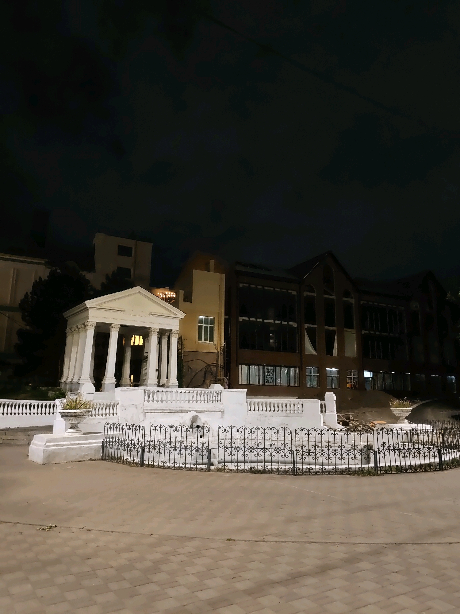 The image depicts a night scene in Rostov-on-Don, Russia, showcasing a white neoclassical portico with Corinthian columns, a white balustrade, and decorative planters. Behind the portico, a dark, multi-story brick building with large, dark windows is visible. The ground in the foreground is paved with light brown rectangular tiles. A black wrought-iron fence with ornate scrollwork encloses a section of the white structure. The sky above is dark, indicating nighttime. There are no people or animals visible in the image.