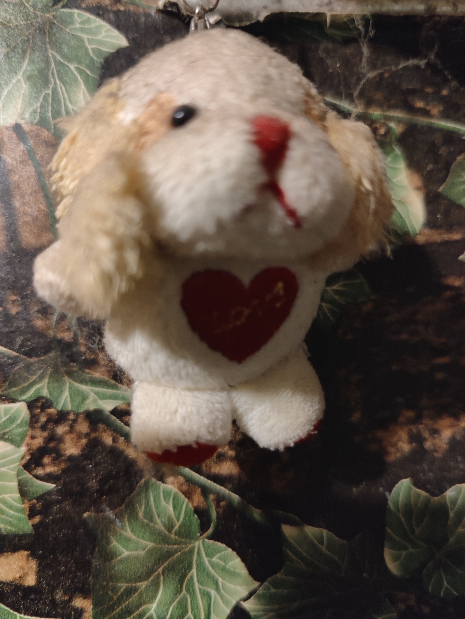 A stuffed toy dog with a red nose and a red heart with the word "LOVE" embroidered on its chest is displayed on a surface with a green leaf pattern. The toy is light brown and white, with floppy ears and black eyes. The background appears to be a dark, textured surface with a repeating pattern of large, green leaves.