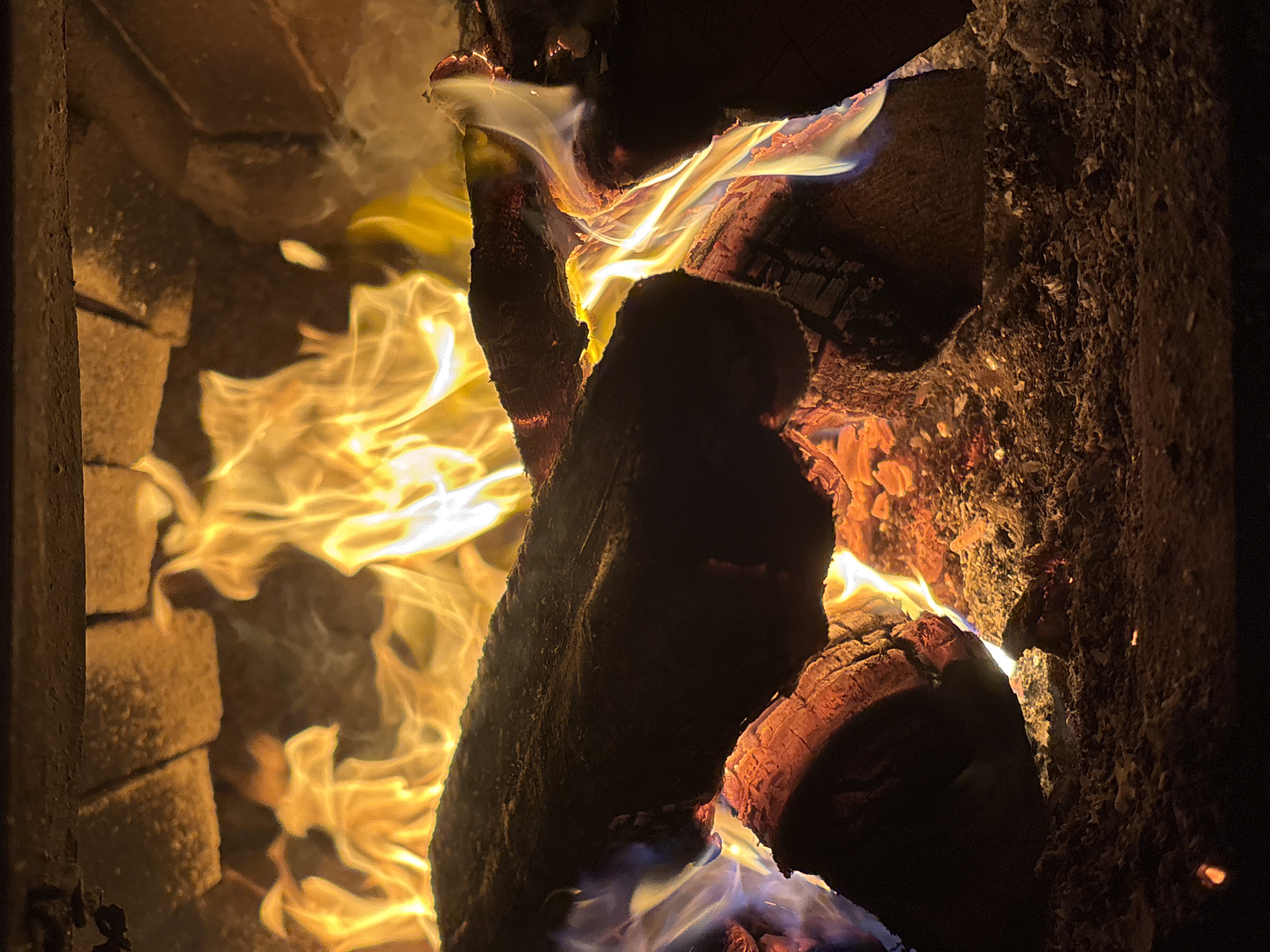 The image depicts a close-up view of a burning fireplace. The scene is dominated by vibrant flames, primarily in shades of yellow and orange, licking at several logs of wood. The logs are dark and appear to be partially charred, with glowing embers visible at their bases. The surrounding environment consists of a brick interior, likely a fireplace or fire pit, with visible ash and debris. There is no indication of any specific location within the city of Курган, Russia. The image focuses solely on the natural phenomenon of a fire. There are no people or animals present in the image.