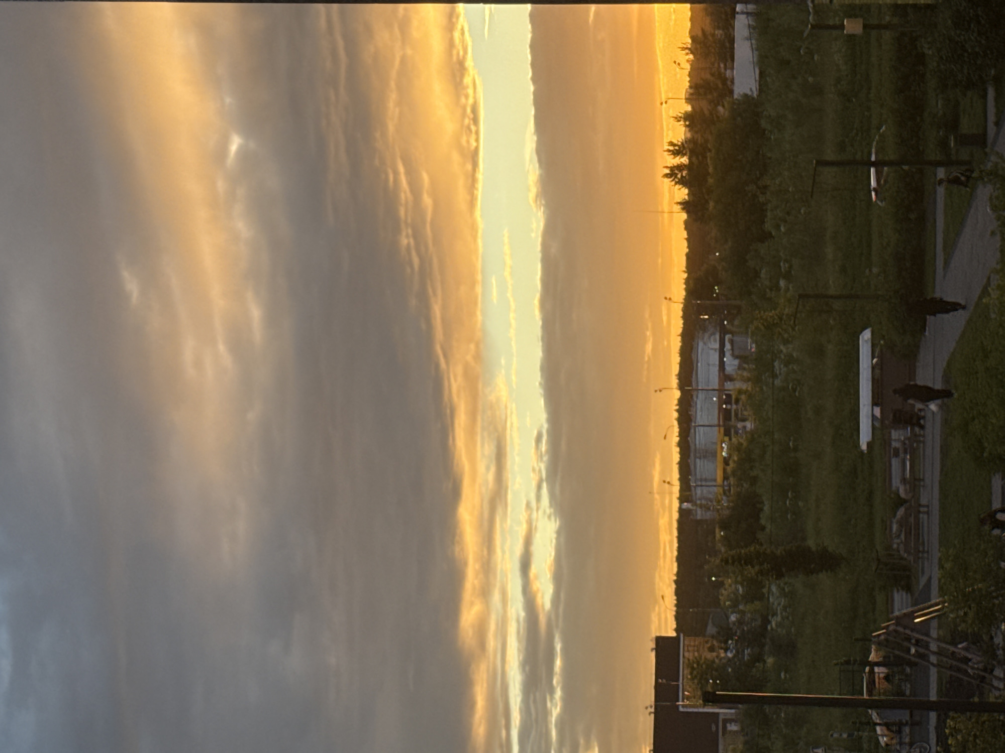 This media file depicts a sky filled with dramatic clouds at sunset. The sky is dominated by shades of orange, yellow, and gray, indicating a sunset or sunrise. The clouds are illuminated by the low sun, creating a striking contrast between light and shadow.
Below the sky, a landscape is visible. The upper portion of the landscape features a treeline silhouetted against the bright sky. The lower portion of the landscape appears to be a mix of grassy areas and possibly some urban or suburban elements like buildings and streetlights. There are lampposts visible in the distance, and a paved walkway with benches can be seen in the foreground on the right side. Some dark shapes on the ground in the foreground might be people or objects, but they are not clearly discernible.
The time of day is clearly sunset or sunrise due to the intense colors in the sky. The weather appears to be partly cloudy, with the clouds contributing to the dramatic visual effect. The overall mood is serene and picturesque, evoked by the beautiful natural light. There is no visible text in the image. The location context is Drozhzhino, Russia.
