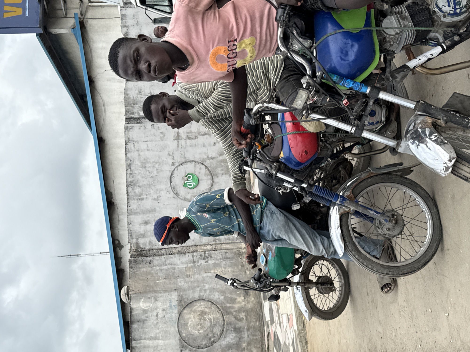 The image shows three young men in Lekki, Nigeria posing with motorcycles. One man wears a pink shirt with "Gucci" text, the second wears a striped shirt, and the third wears a patterned blue shirt and a dark blue cap with orange trim. They are positioned behind a row of three motorcycles with various colored fuel tanks. A white van is partially visible in the background. A blue and white building is on the left. The background wall displays the "glo" logo. The ground is paved.