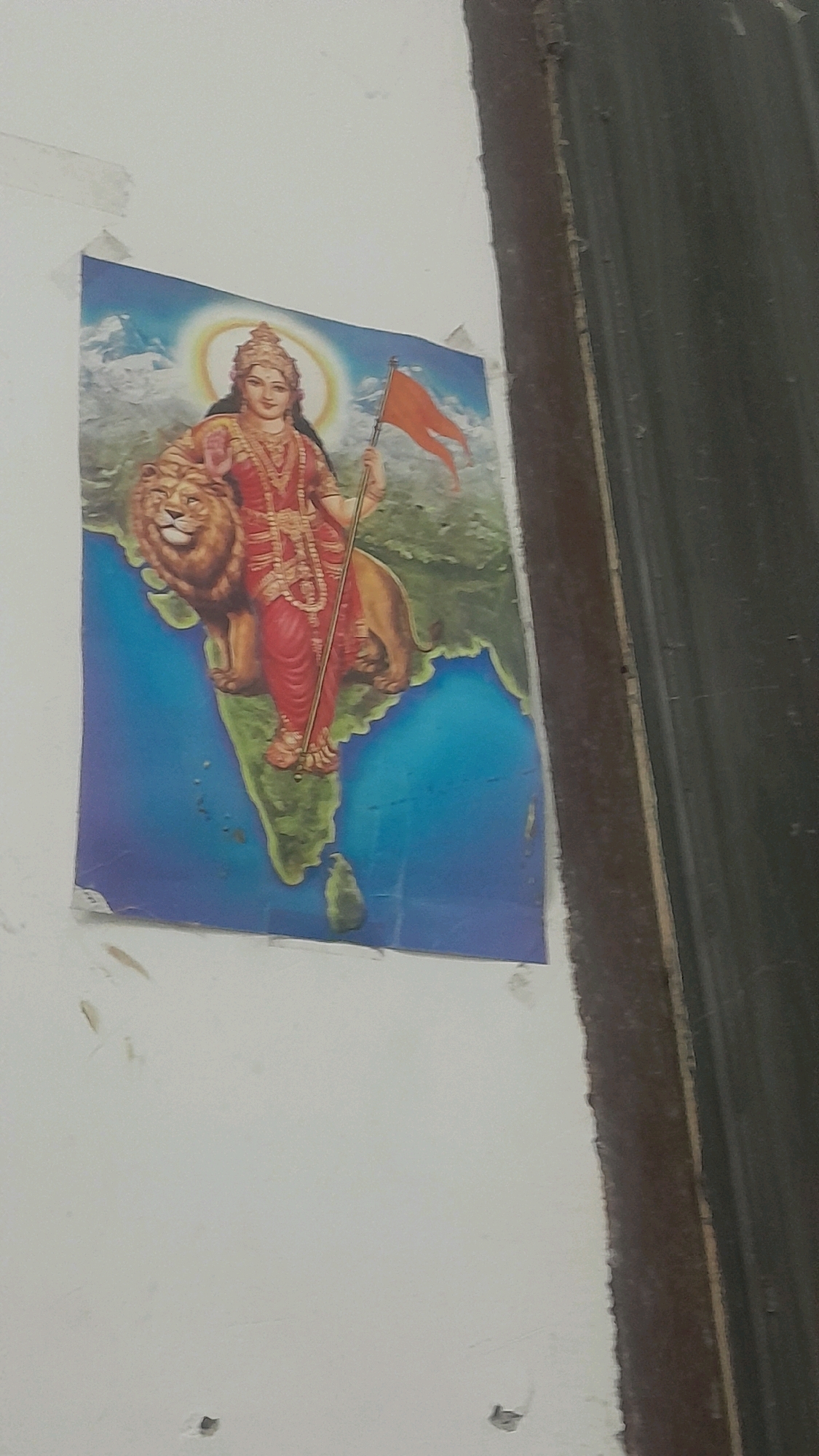 The image shows a religious poster on a white wall, likely in Agra, India. The poster depicts a female deity, possibly Durga, riding a lion and holding an orange flag. The background of the poster appears to be a map of India. The wall is textured and shows some signs of wear, with a dark, corrugated material bordering the right side of the image. The lighting is dim, suggesting the image was taken indoors.